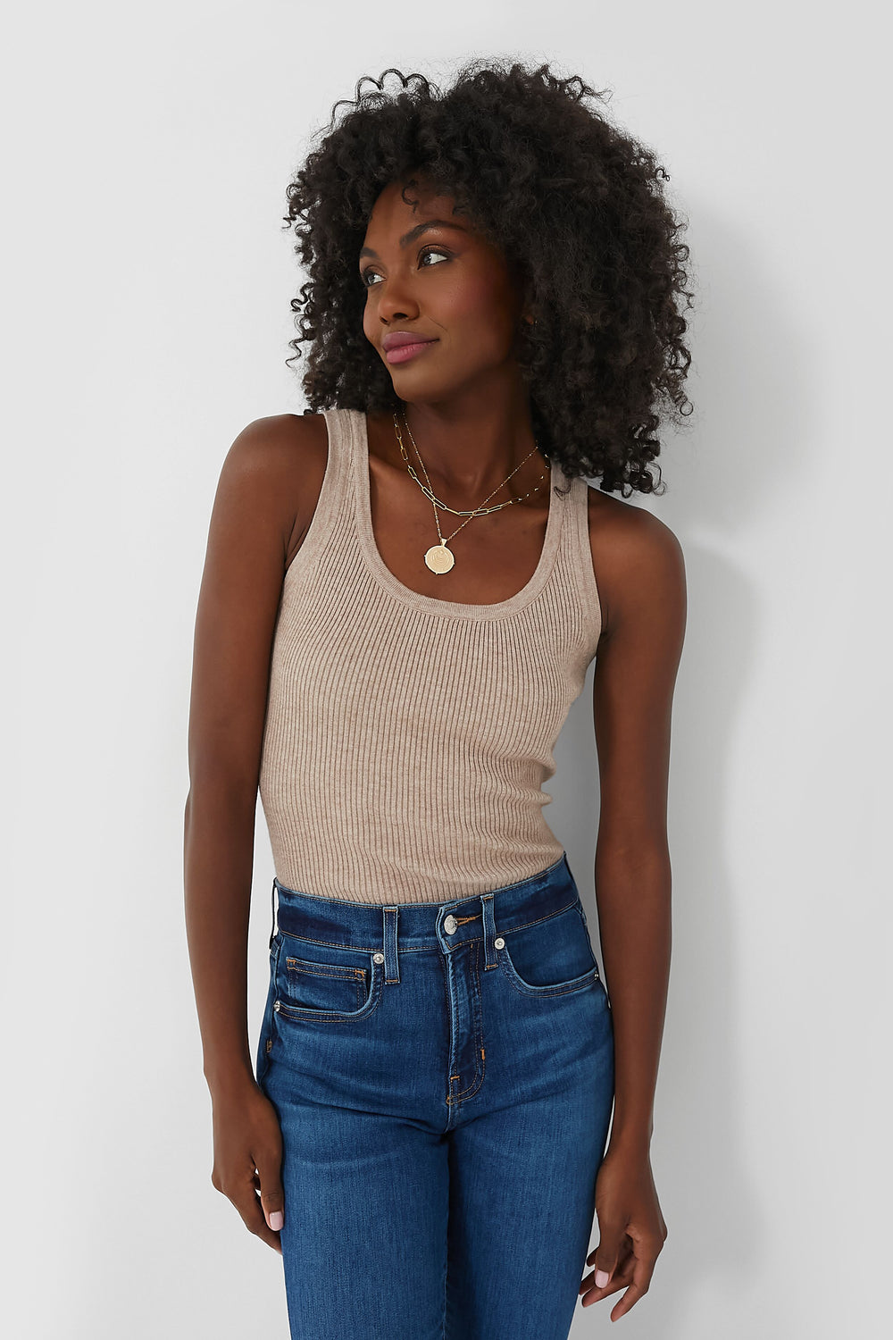 A woman with curly hair wears a Tuckernuck beige slim fit ribbed tank top and blue jeans, embodying the effortless style of elevated basics.