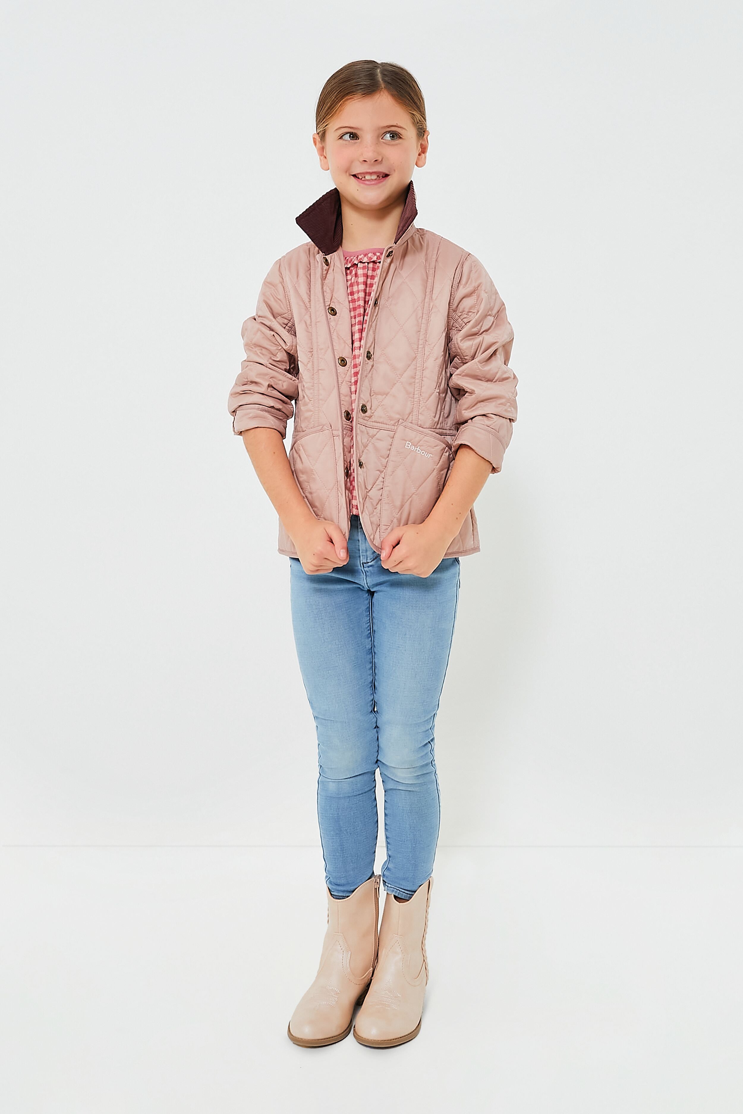 A young girl wears a light pink Barbour girls quilted jacket over a red checkered shirt, paired with blue jeans and beige boots.