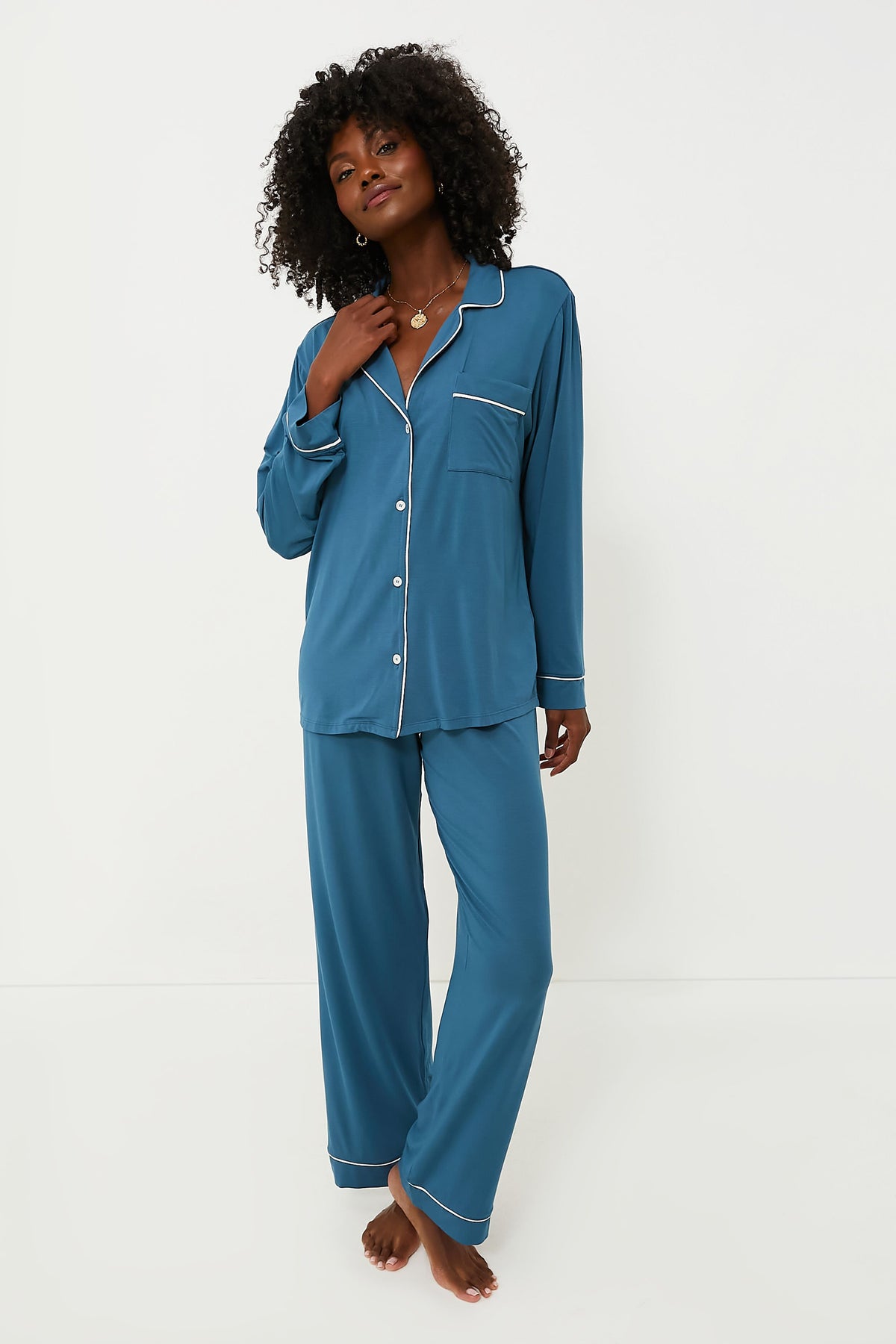 A woman stands barefoot in Eberjey’s blue long-sleeve button-up pajama set with white piping, crafted from plant-based TENCEL™ Modal for sustainable comfort.