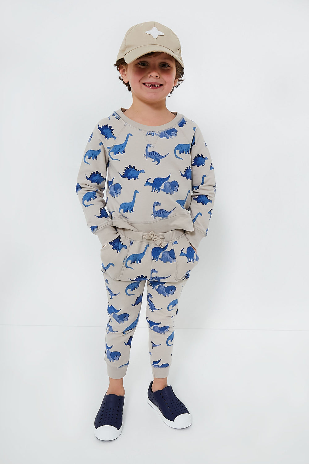 A young child models an Angel Dear beige jogger set with blue dinosaur prints, paired with a beige cap and blue slip-on shoes—stylish organic cotton baby clothing.