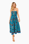 A woman wears the Ulla Johnson Lagoon Ella Dress, a blue floral-print, 100% cotton halter-neck midi with a gathered bodice and flowy skirt, styled with tan lace-up sandals.