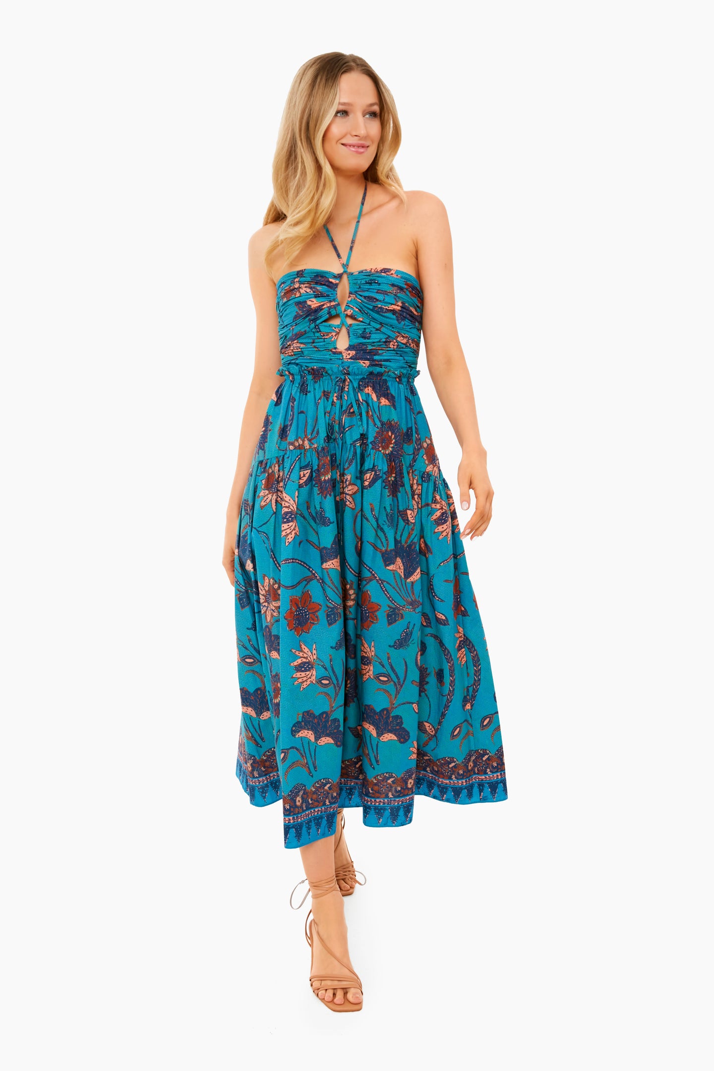 A woman wears the Ulla Johnson Lagoon Ella Dress, a blue floral-print, 100% cotton halter-neck midi with a gathered bodice and flowy skirt, styled with tan lace-up sandals.