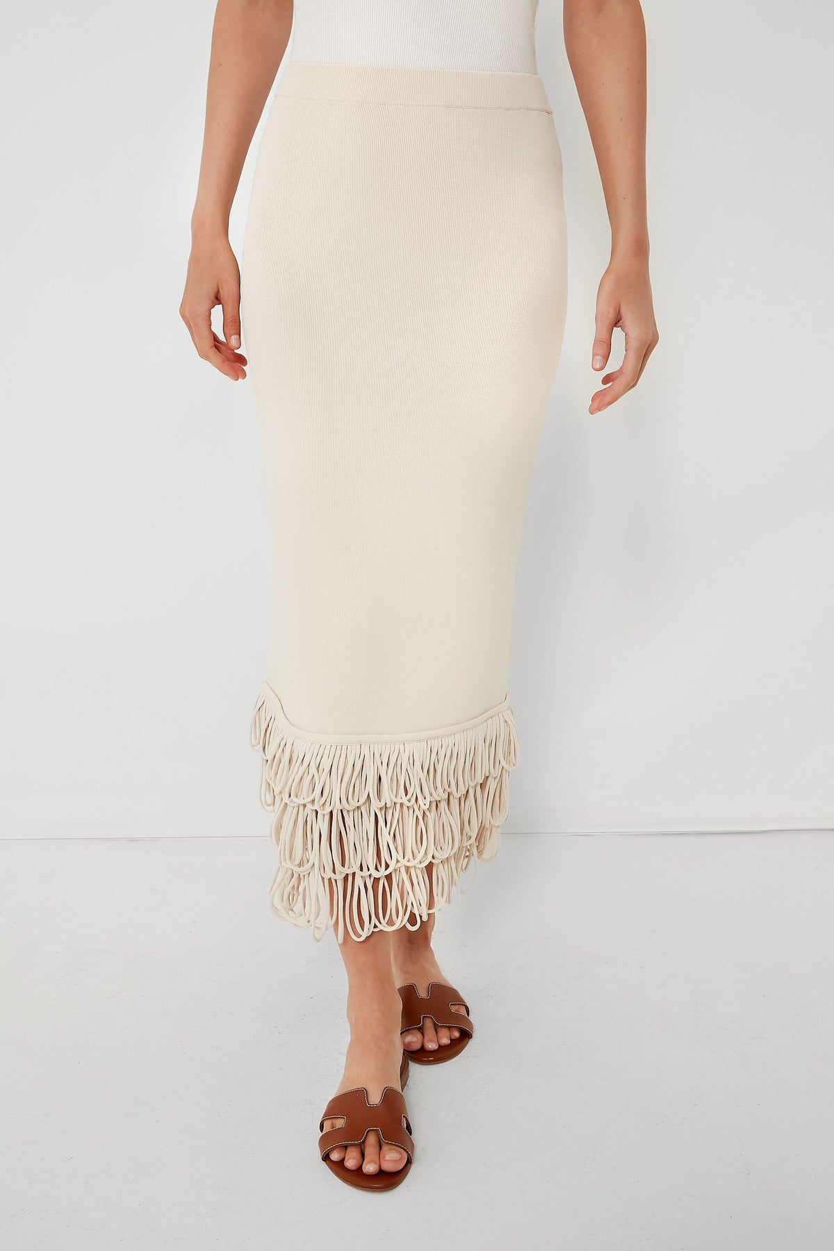 Wearing a Simon Miller Ecru Sol Skirt with a fringe hem and brown flat sandals, this look highlights the cream-colored midi skirt’s stylish details.
