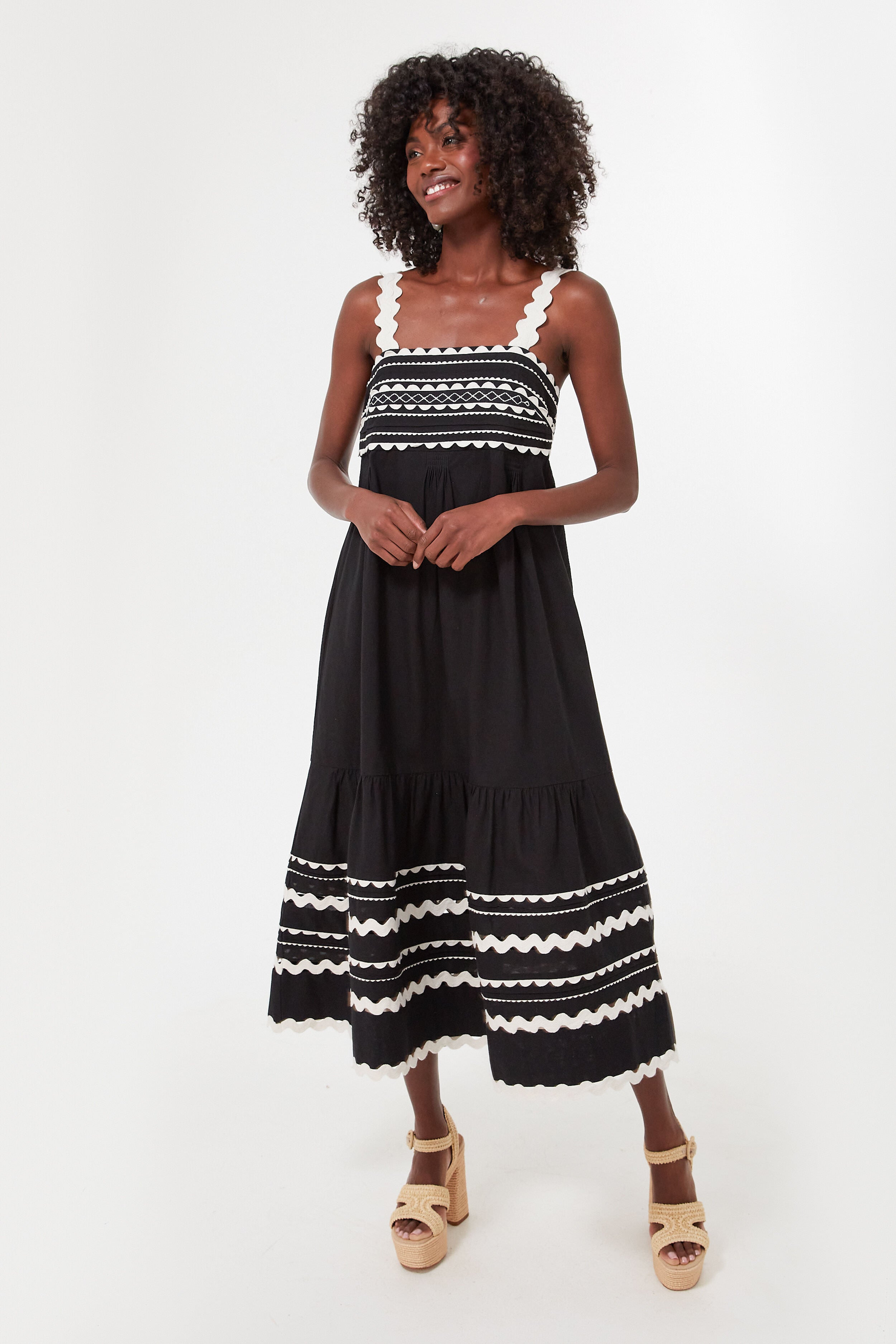 A woman smiles while wearing the Sea New York Ryleigh Dress, a sleeveless black midi with white zigzag trim, paired with beige platform sandals.