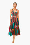 A woman wears the Ulla Johnson Varda Dress, a sleeveless, multicolored patterned hand-loomed dress with a fitted bodice and pleated skirt, styled with strappy sandals and green earrings.
