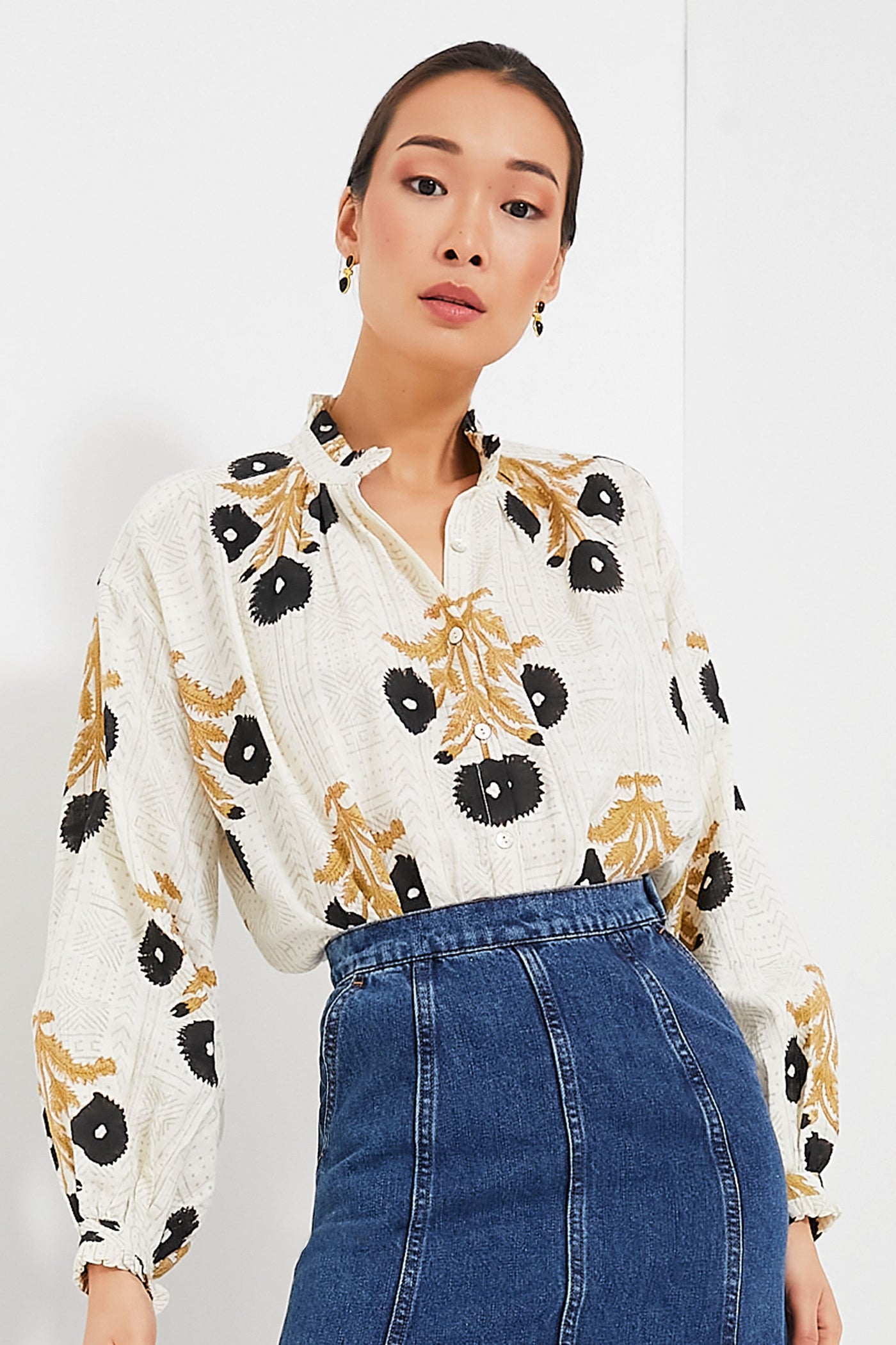 A woman wears an Alix of Bohemia boho-chic blouse featuring black and gold floral patterns with a ruffle collar, styled with high-waisted blue jeans.