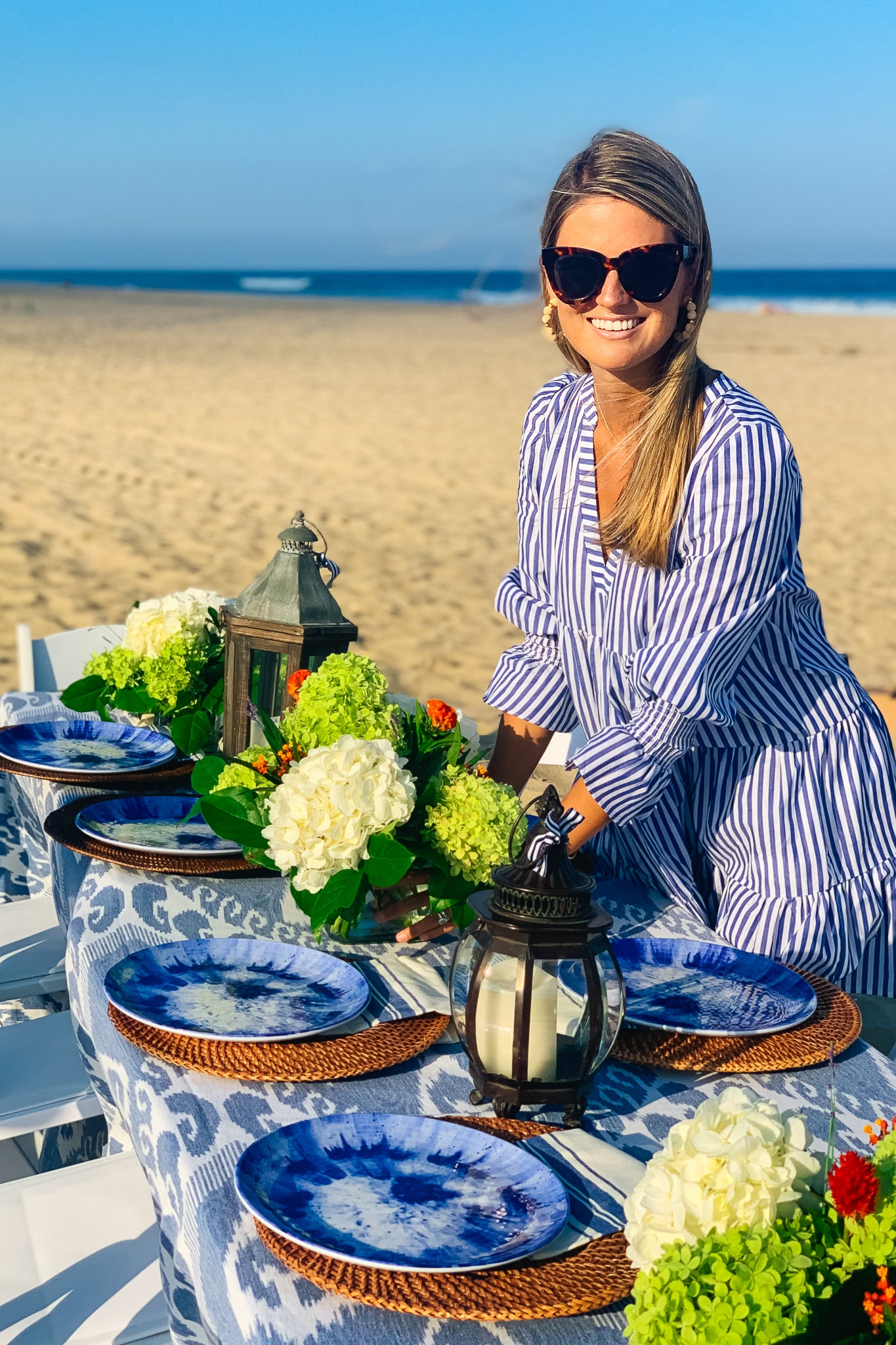 A woman in sunglasses and a Pomander Place blue and white striped dress stands by a beach table set with blue plates, lanterns, and flowers under a clear sky.
