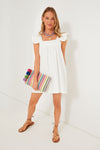 A woman in a white Pomander Place cotton mini dress with flutter sleeves and brown sandals holds a striped clutch, smiling at the camera.