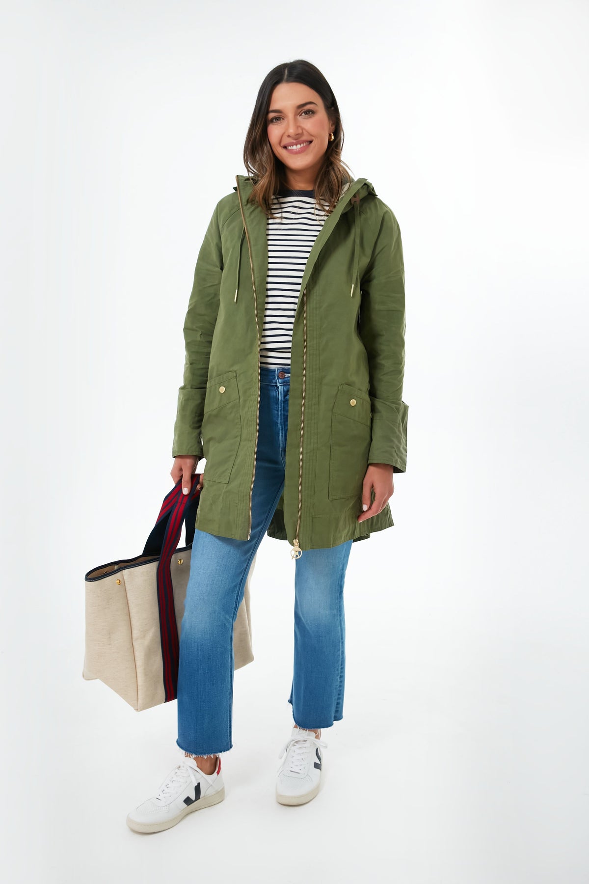 A woman smiles wearing a green Barbour showerproof parka, striped shirt, blue jeans, and white sneakers, holding a beige tote bag with dark handles.