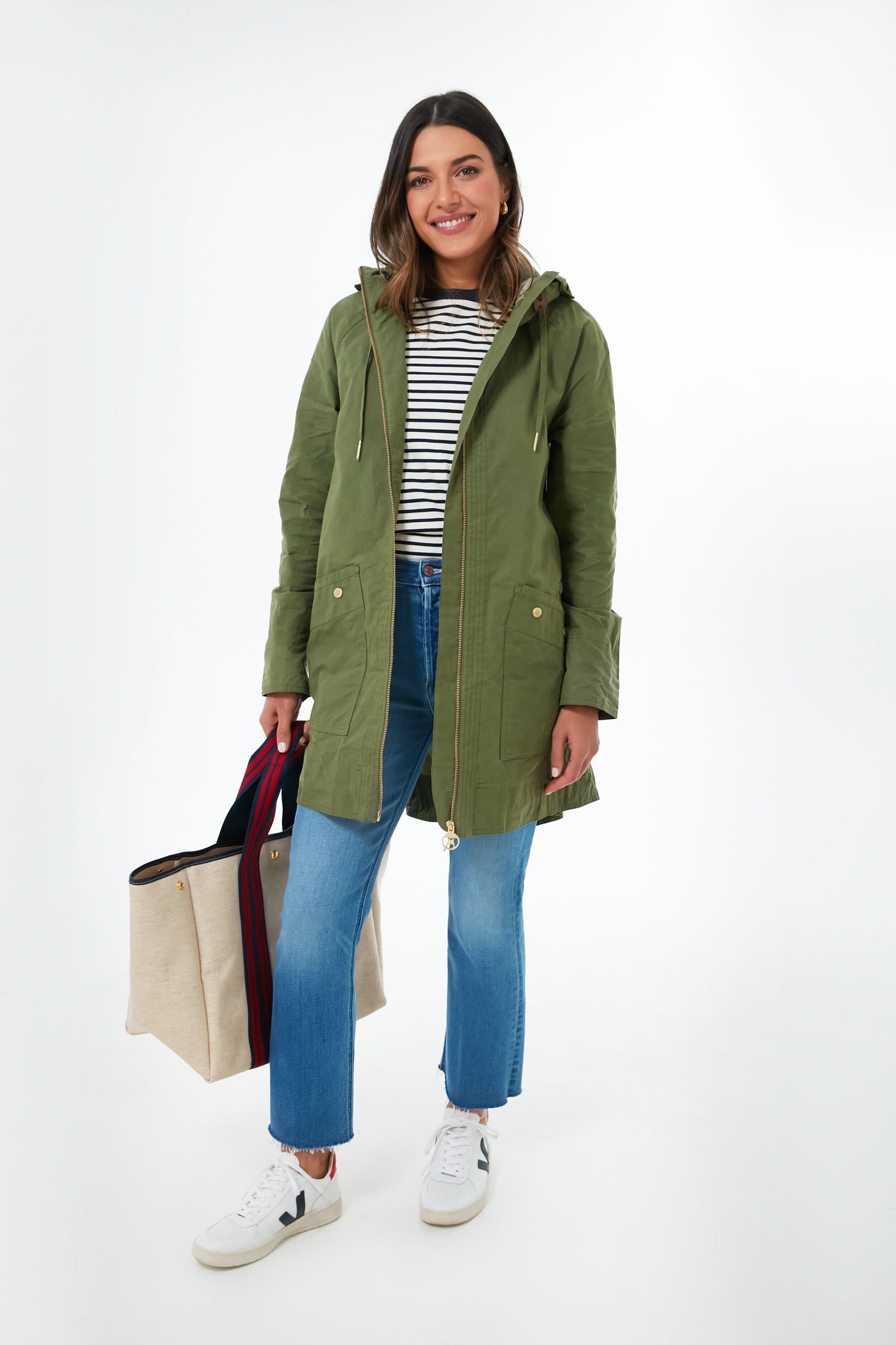 A woman smiles wearing a green Barbour showerproof parka, striped shirt, blue jeans, and white sneakers, holding a beige tote bag with dark handles.