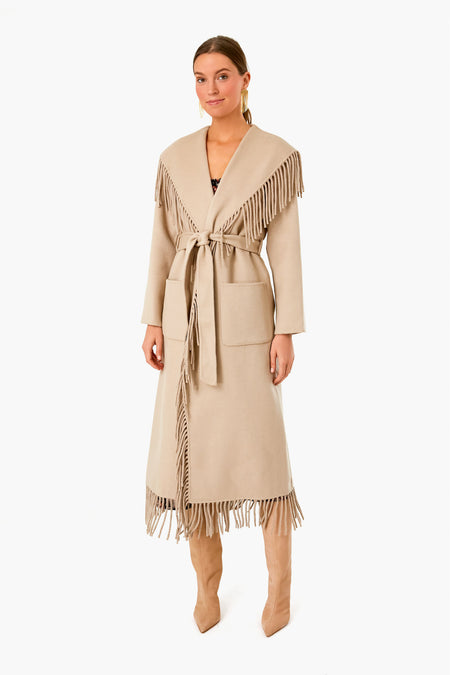 A woman wears a luxury outerwear piece—a long beige wool coat by SIMKHAI, featuring fringe on the collar and hem, a tie belt at the waist, and tan knee-high boots.