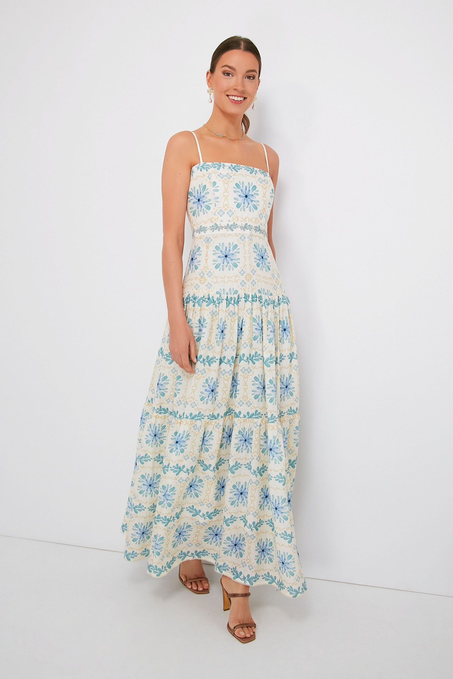 A woman smiles brightly wearing a sleeveless, ankle-length tropical floral dress in blue and yellow patterns by AGUA BY AGUA BENDITA.