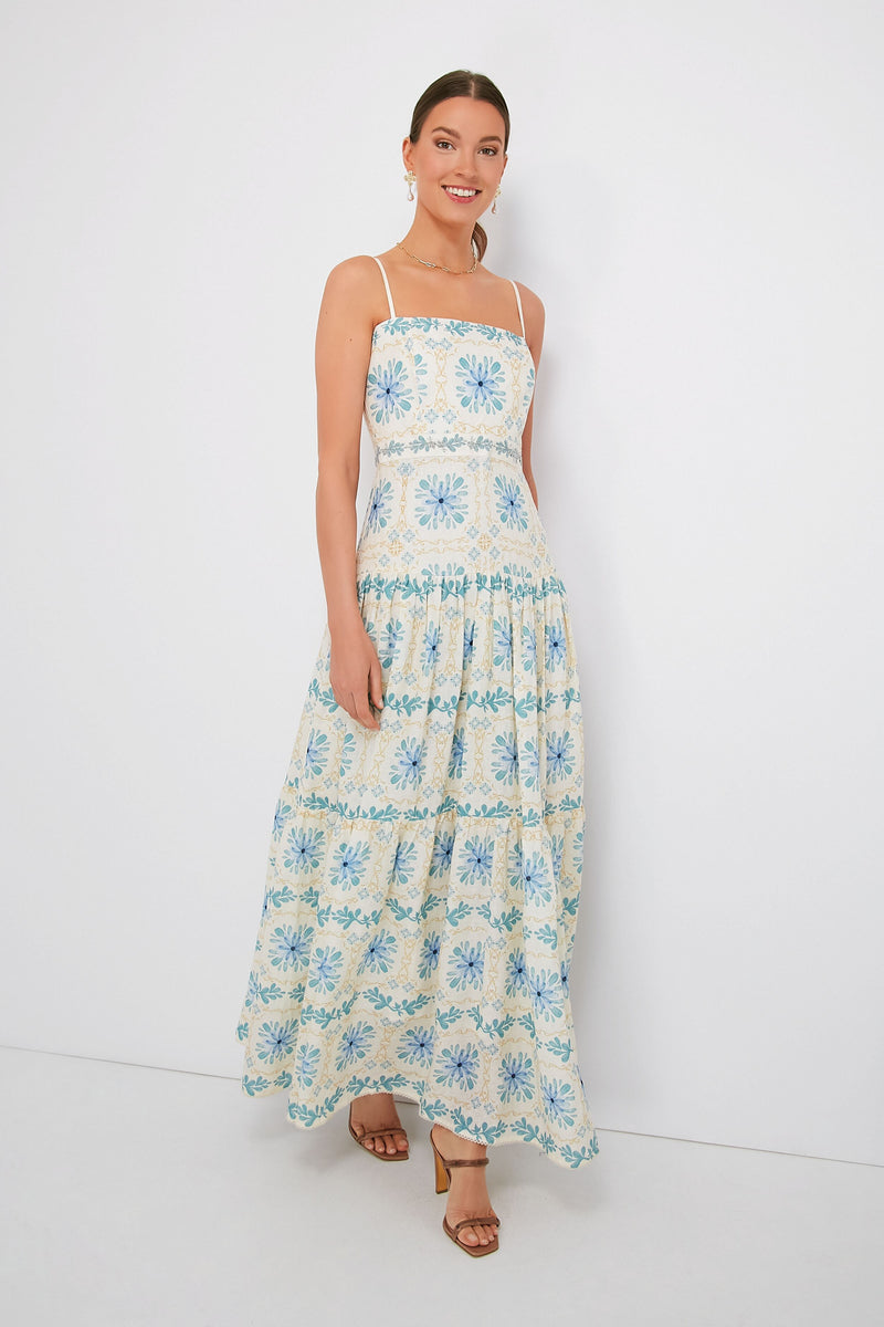 A woman smiles brightly wearing a sleeveless, ankle-length tropical floral dress in blue and yellow patterns by AGUA BY AGUA BENDITA.