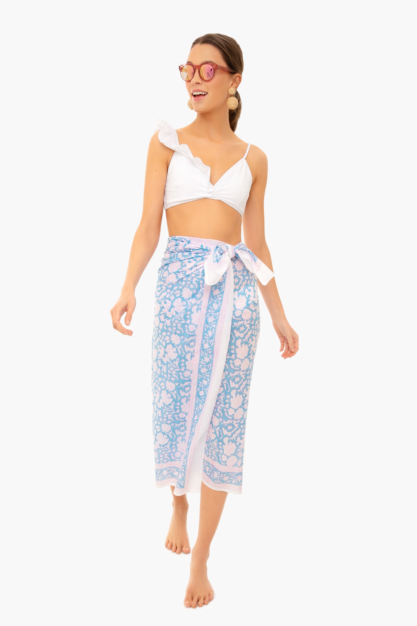 A woman in a white bikini top, pink sunglasses, and an SZ Blockprints light blue and pink hand block printed cotton Indian sarong as a beach coverup stands barefoot.