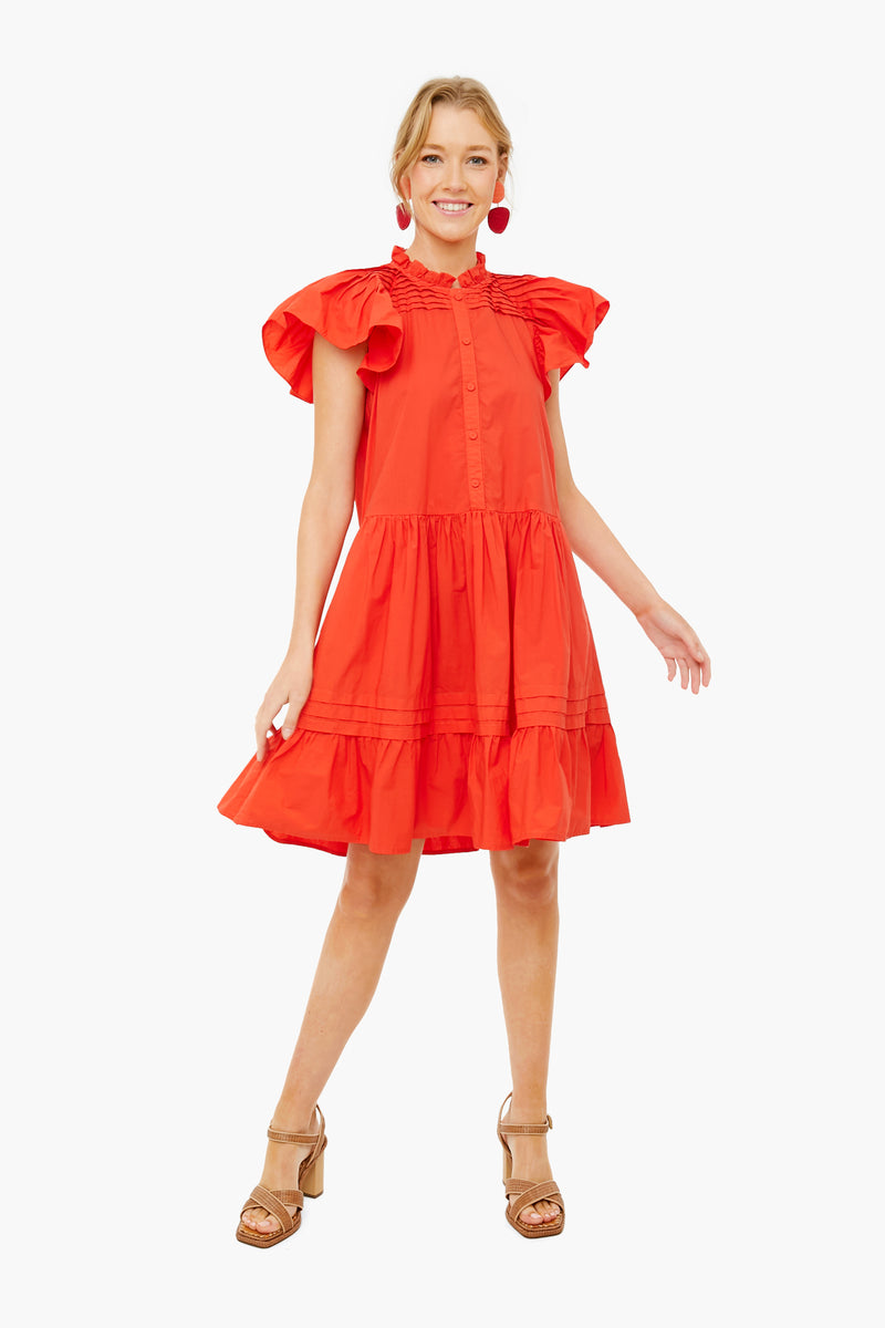 A woman wears a bright red Sea New York cotton mini dress with pleated details, tiered skirt, short sleeves, and button front, paired with tan heels and red earrings.