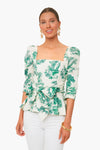 A woman styles the Cara Cara Lauren Top—a white blouse with green forest botanical print, square neckline, puffed sleeves, and waist tie—with white pants, gold earrings, and bangles.