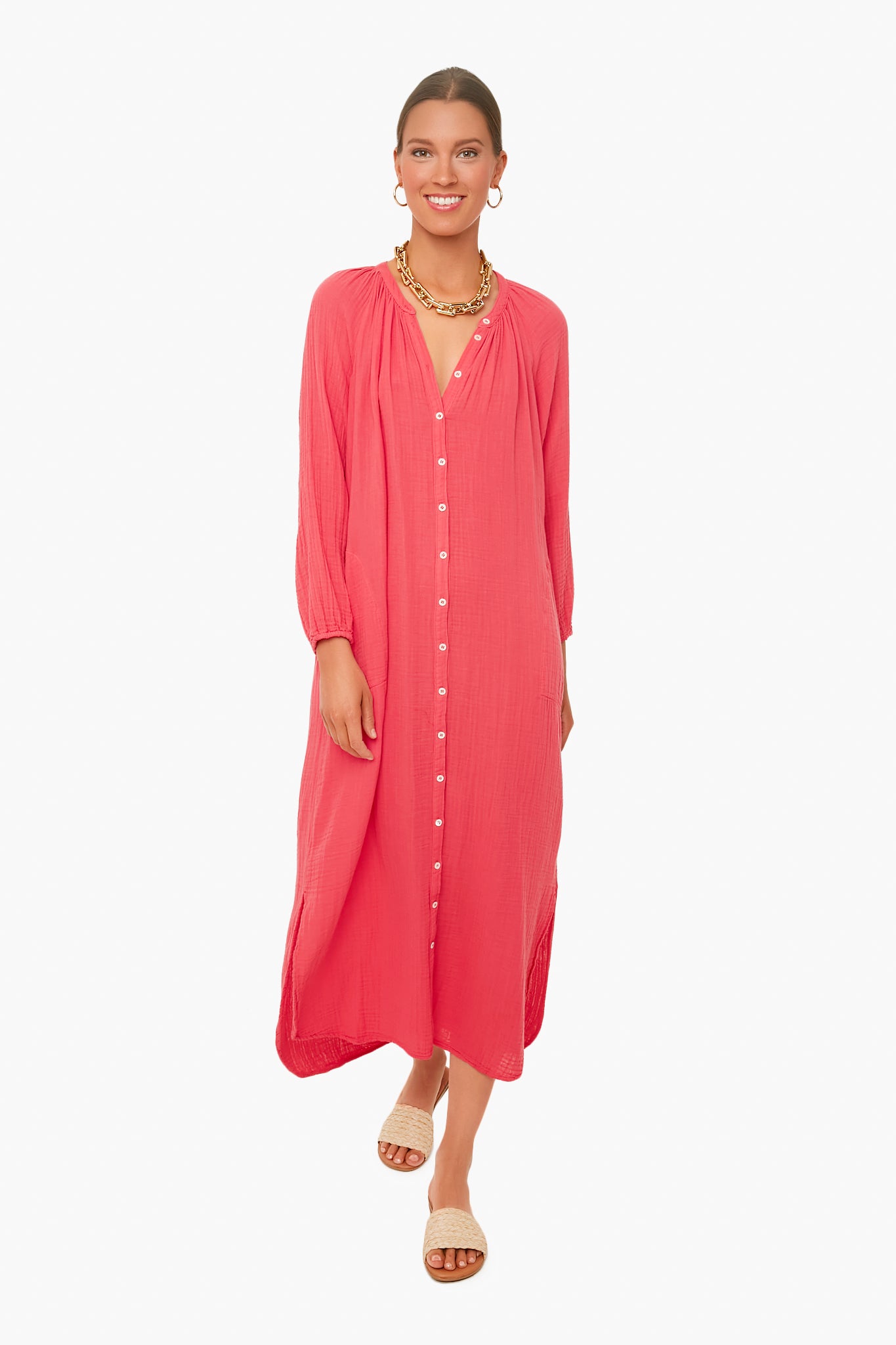 A woman wears a pink XiRENA cotton gauze v-neck summer dress, styled with a gold necklace, hoop earrings, and open-toe sandals as she stands confidently.
