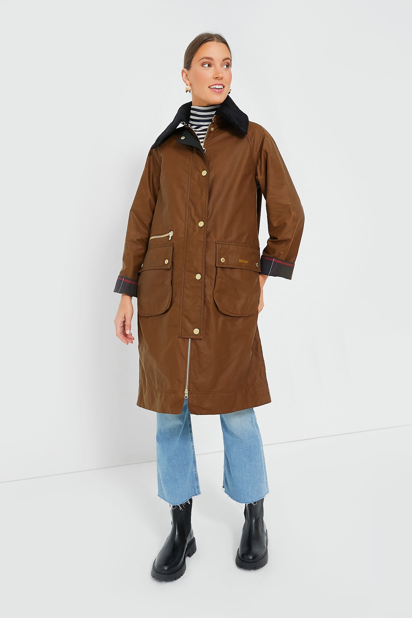 A woman stands confidently in a Barbour brown waxed cotton coat with gold buttons, blue jeans, black boots, and a striped turtleneck.