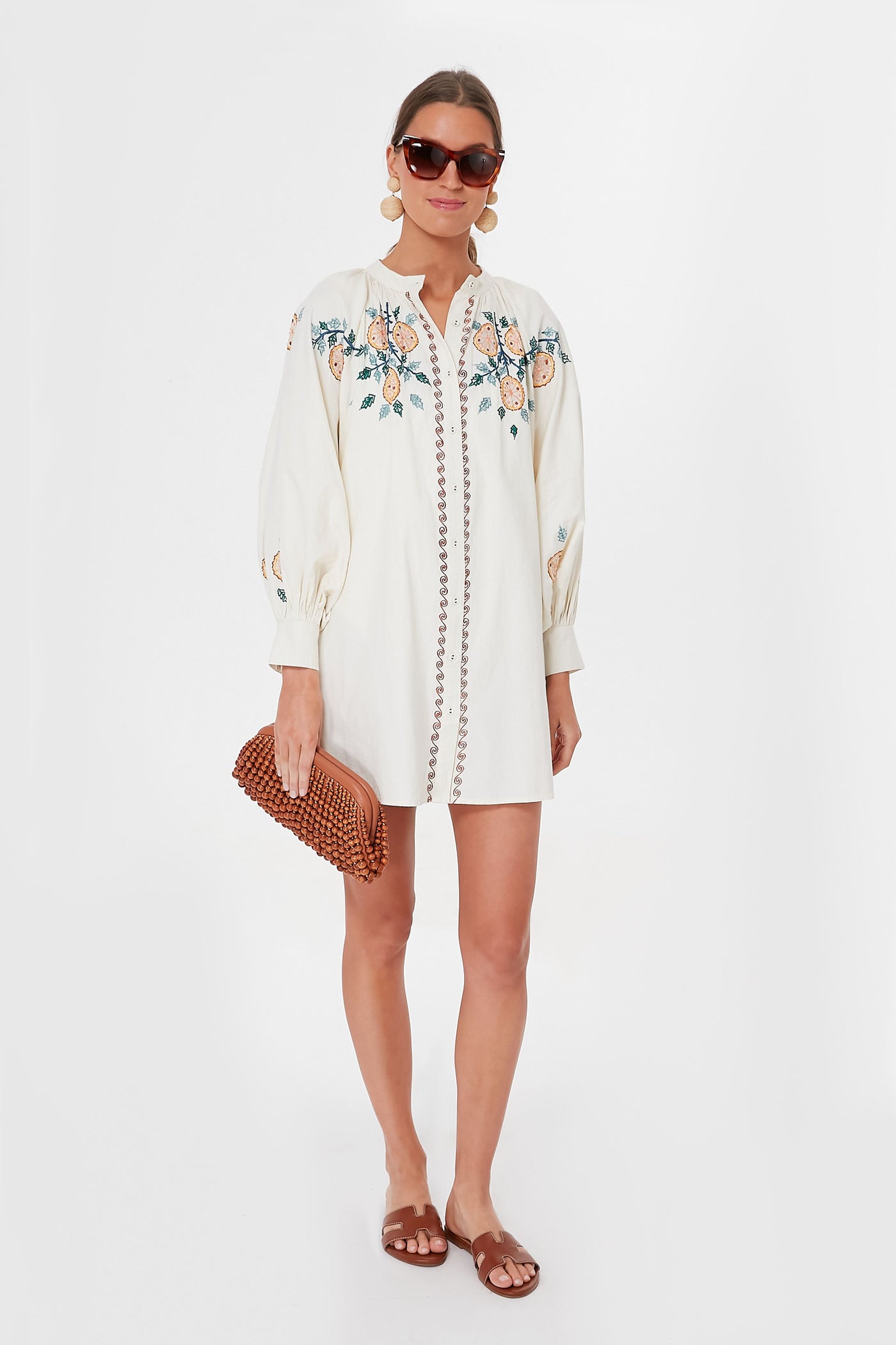 Woman wearing a white embroidered Carolina K. Gardenia Augustina Dress with long sleeves, brown sandals, large earrings, sunglasses, and holding a woven clutch.