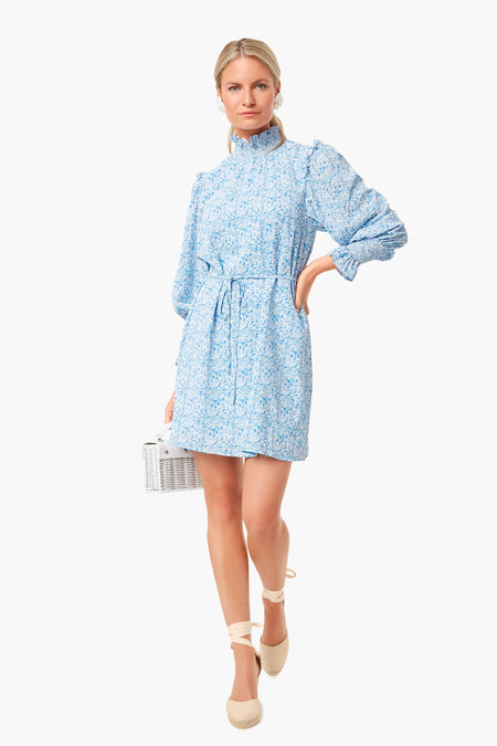 A woman wears the Pomander Place Balou Mini Dress, a blue and white floral long-sleeve dress with a ruffled collar and tie waist, styled with espadrille shoes and a small white handbag.