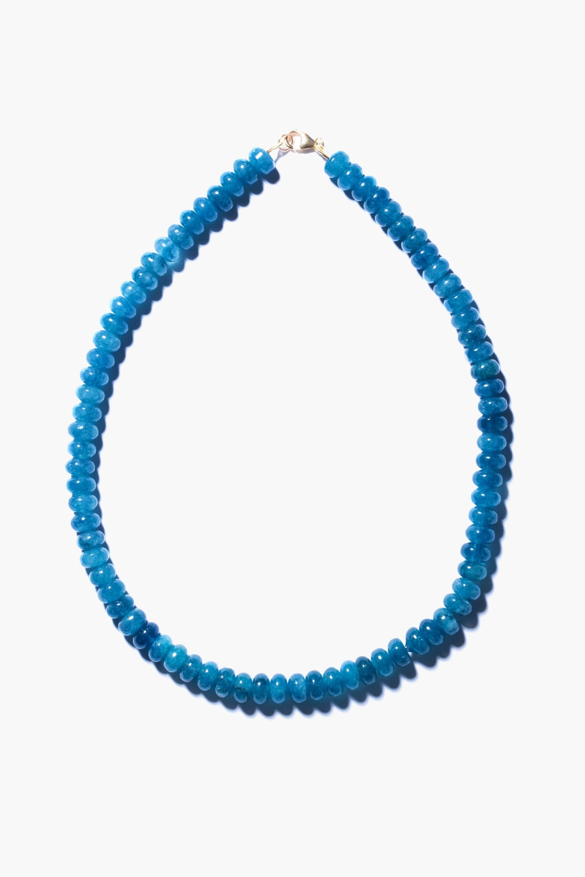 This handmade angelite necklace by Theodosia features round, bright blue beads with a small gold clasp, elegantly arranged in an oval shape—a stunning piece of jewelry.