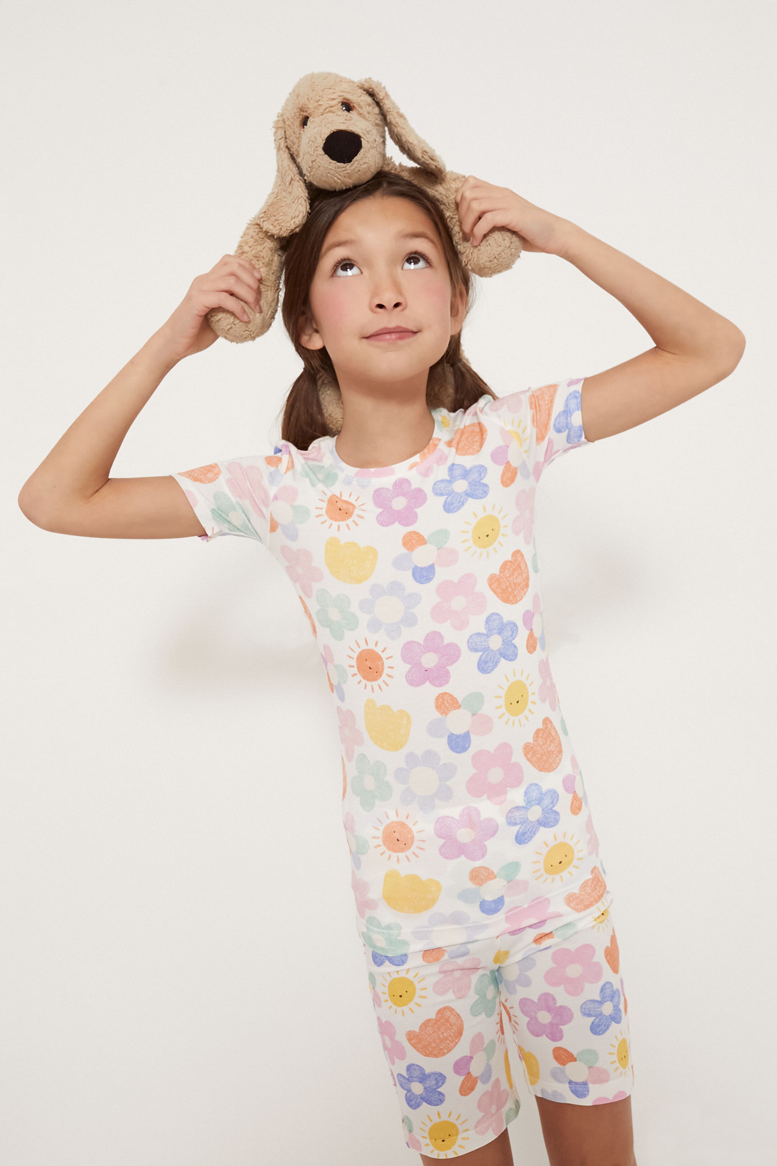 A young girl wearing Angel Dear bamboo baby pajamas with floral prints holds a stuffed dog on her head and looks upward.