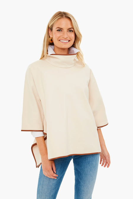 A woman stands smiling in a loose, light beige Tuckernuck Piper Poncho with brown leather trim, blue jeans, and gold hoop earrings.