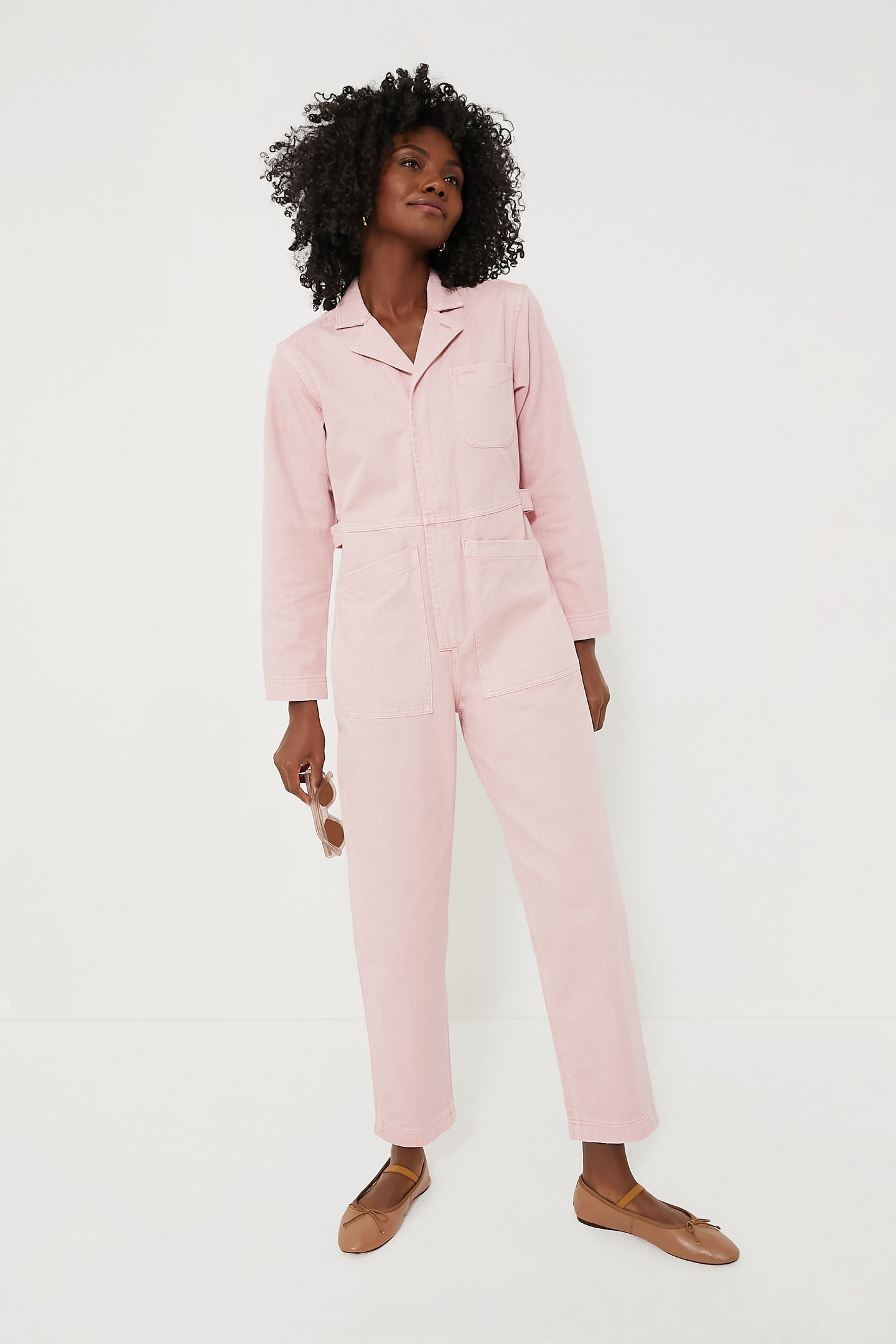 Someone wears a light pink Alex Mill cotton herringbone jumpsuit with brown flats, holding sunglasses in one hand.