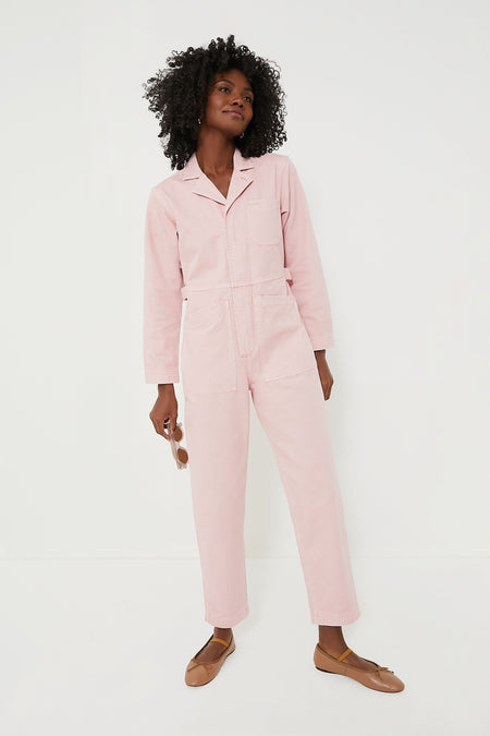 Someone wears a light pink Alex Mill cotton herringbone jumpsuit with brown flats, holding sunglasses in one hand.