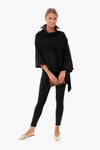 A woman wears a black 100% acrylic layering poncho from Pomander Place, styled with black leggings, gold flats, and a brown clutch.