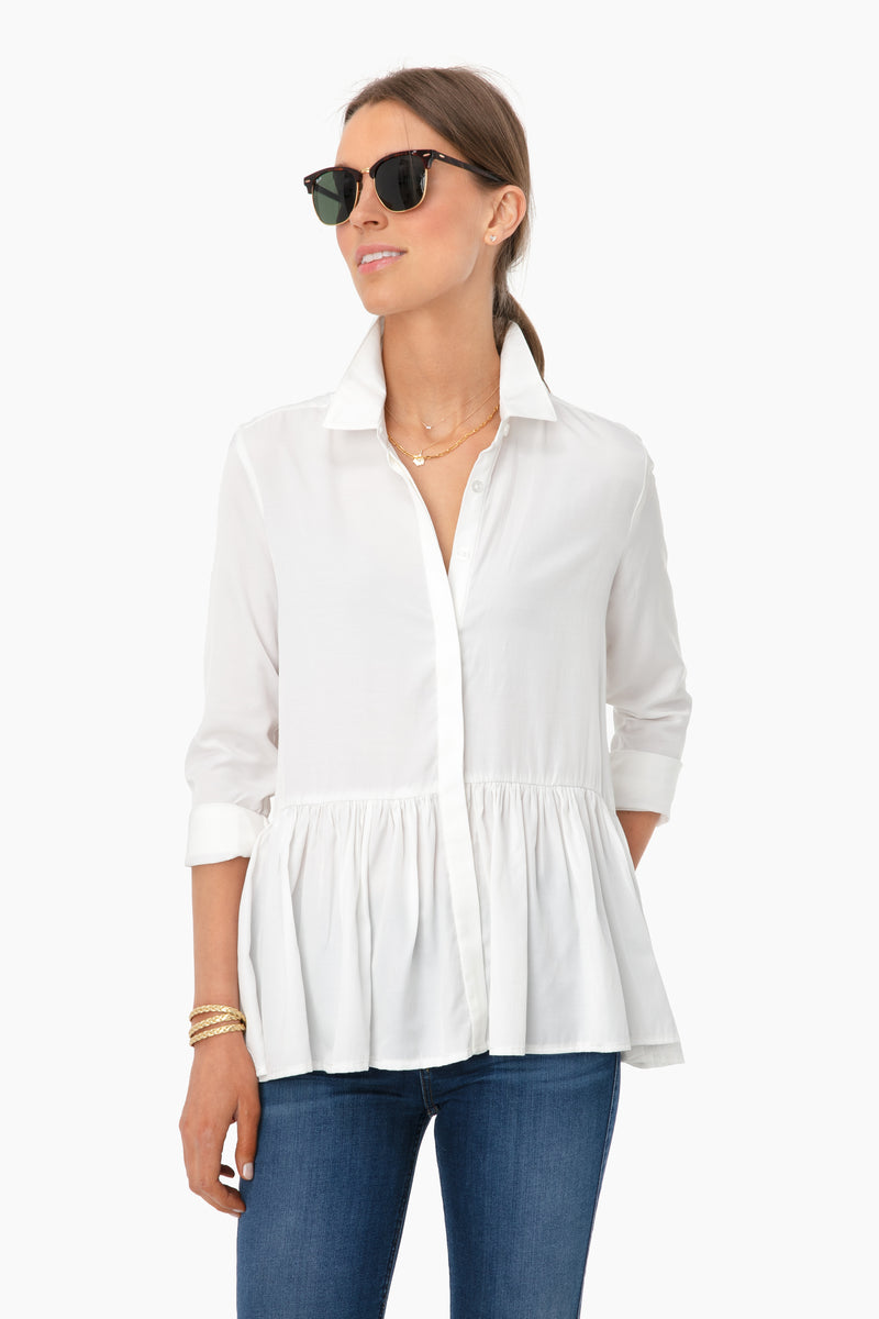 A woman confidently poses in Tuckernuck sunglasses, a white button-down blouse with a ruffled peplum collar, blue jeans, and bracelets.