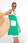 A woman smiles at the camera in a Tuckernuck Kelly Dress with green and white color-blocking and front pocket, holding a wide-brimmed straw hat and wearing white cat-eye sunglasses.