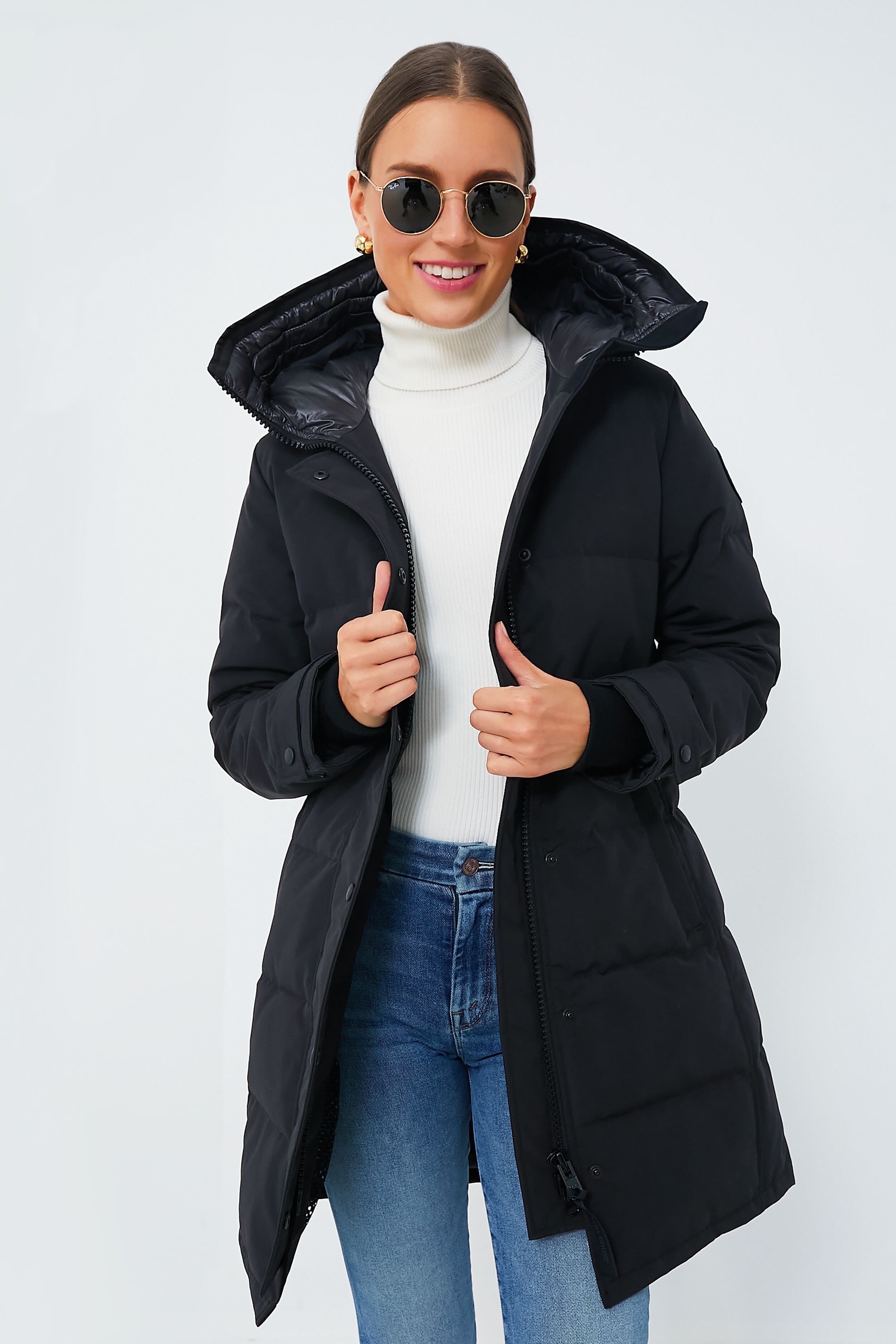 A woman in sunglasses, a white turtleneck, blue jeans, and a black quilted Canada Goose parka stands smiling at the camera.
