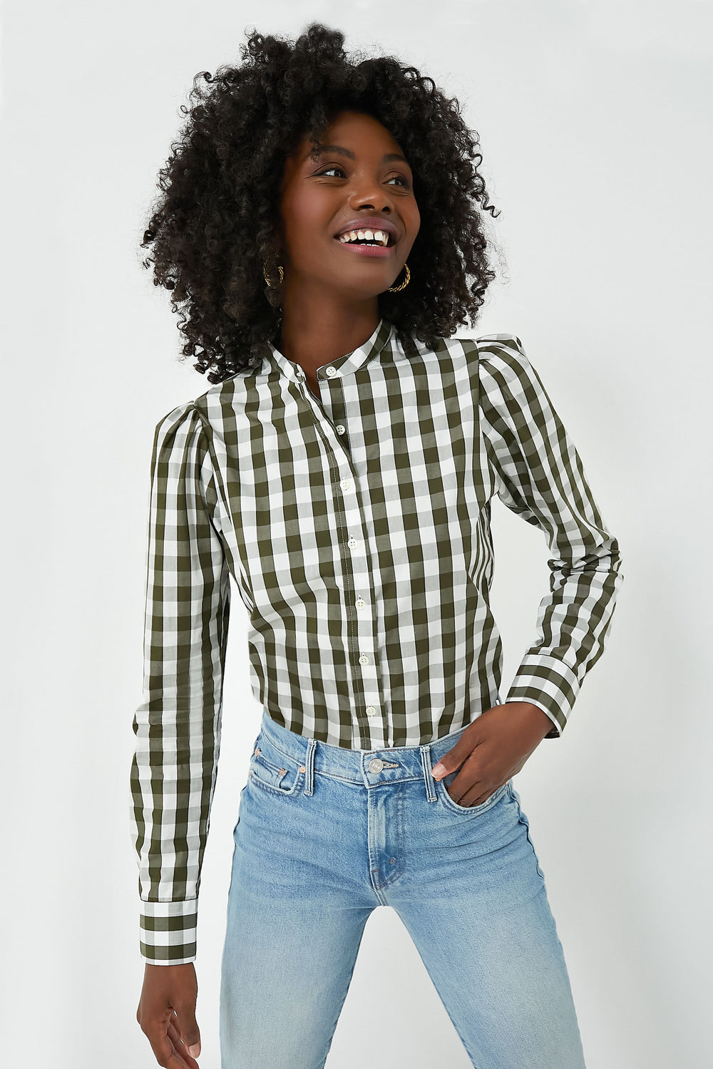 Wearing The Shirt by Rochelle Behrens, a person in a green and white checkered cotton shirt and light blue jeans stands, smiling and looking to the side.