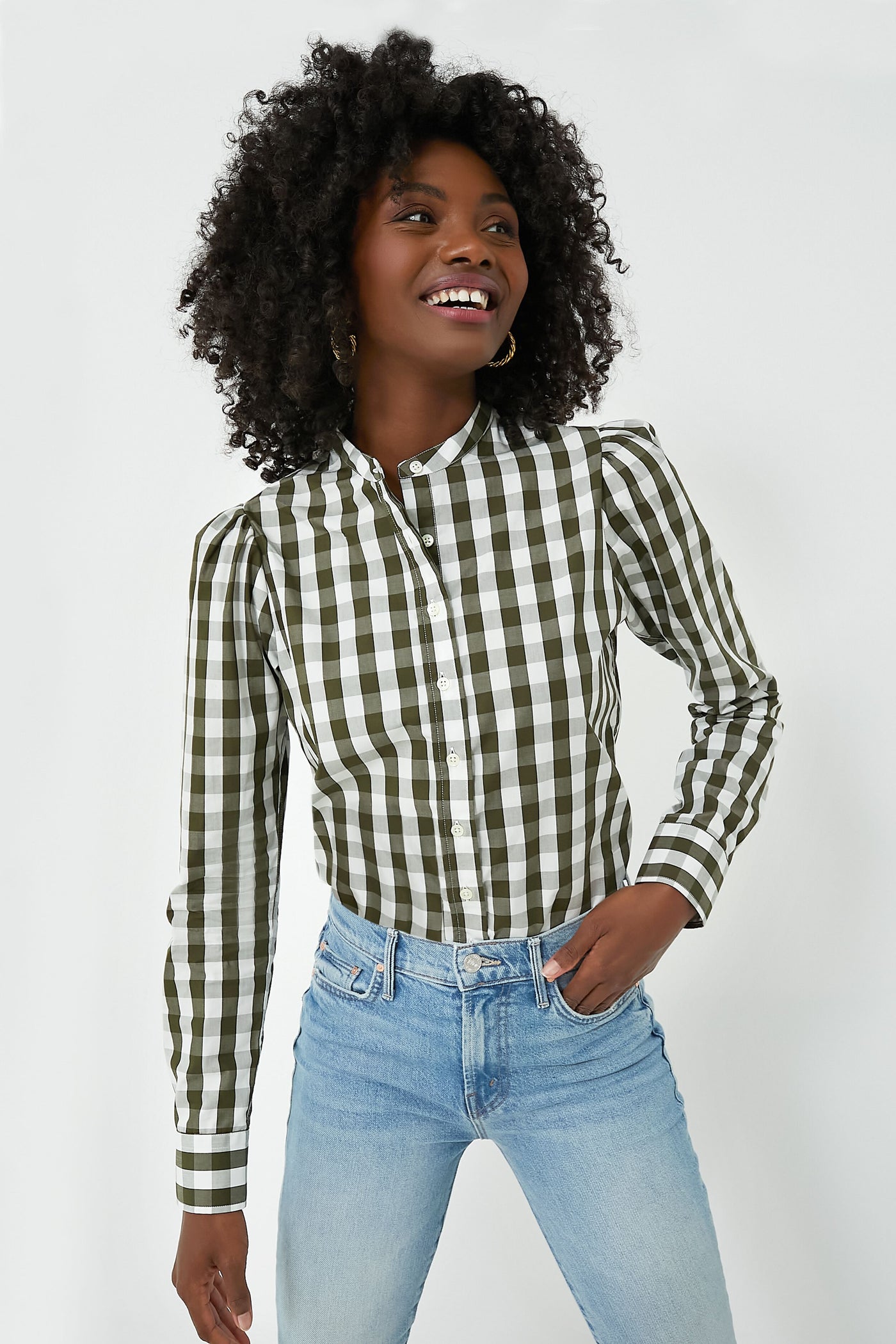 Wearing The Shirt by Rochelle Behrens, a person in a green and white checkered cotton shirt and light blue jeans stands, smiling and looking to the side.