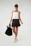 A woman in Tnuck Sport’s white racerback quick-dry tank, black pleated skirt, and visor holds a black bag and tennis racket.