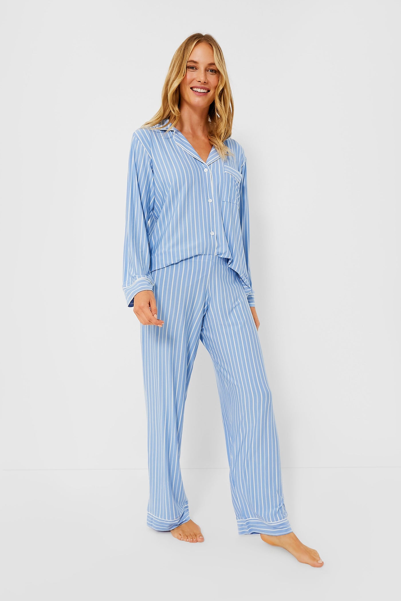 A woman stands barefoot in an Eberjey light blue and white vertically striped TENCEL modal pajama set with a button-up shirt and pants, highlighting comfortable, sustainable sleepwear.