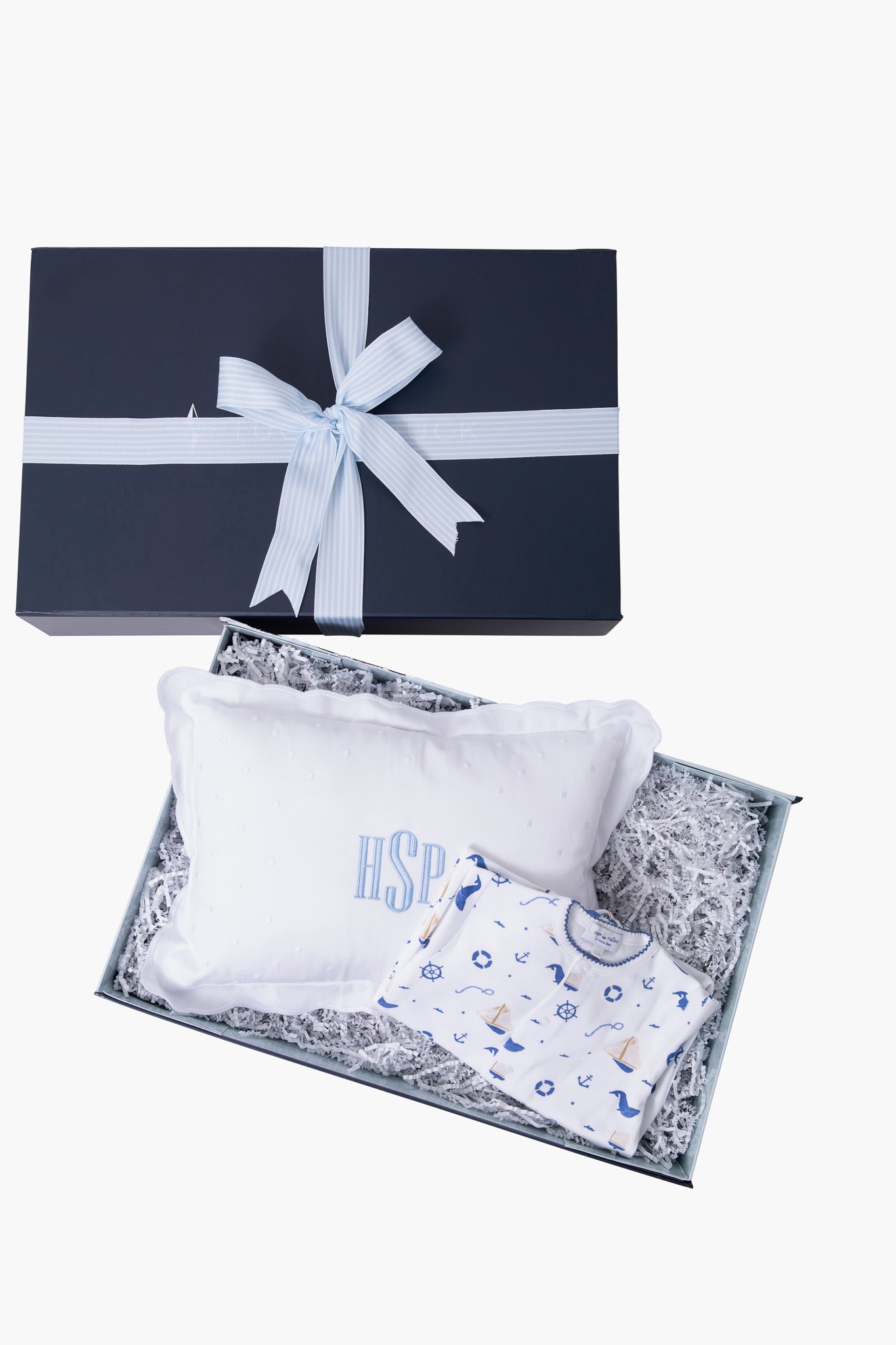 Tuckernuck Tots gift box with a blue ribbon includes a white pillow with blue monogrammed letters and a white baby outfit featuring a blue and yellow pattern, all arranged on shredded paper—ideal for baby gifts or nautical-themed presents.
