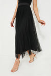 A woman wears a black sleeveless top and the Tuckernuck Black Tulle Kirsten Skirt with a sheer overlay, paired with strappy nude heels, posing confidently.