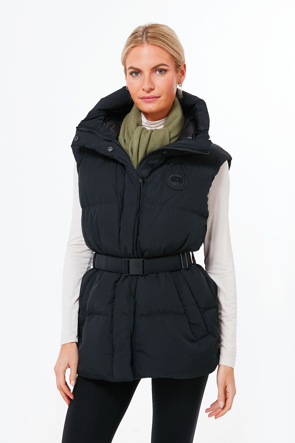 A woman wears a black Canada Goose water-repellent puffer vest over a white long-sleeve top and green scarf, confidently showcasing the brand’s signature outerwear style.