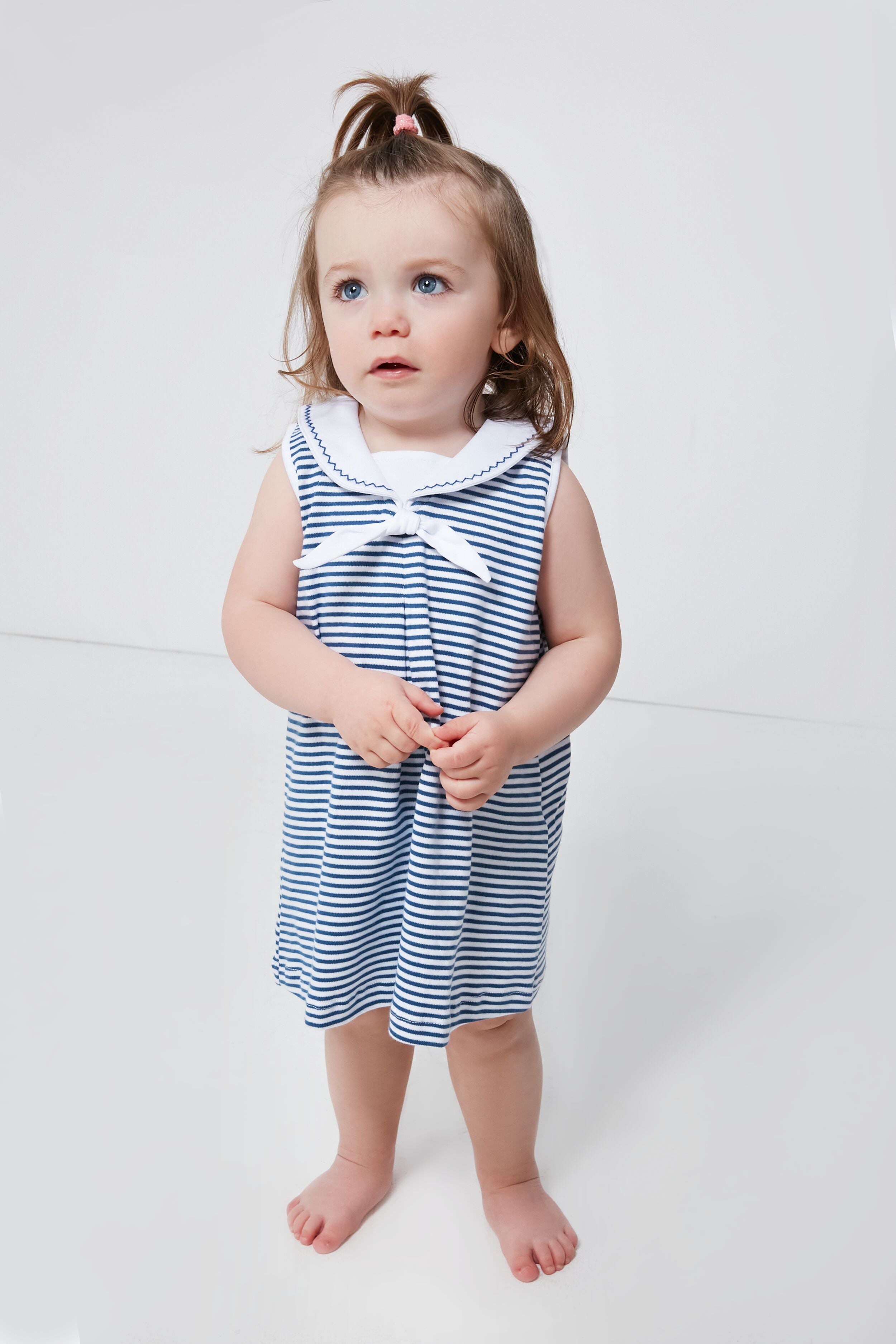 A young child with blue eyes and brown hair wears a sleeveless, blue-and-white striped Pima cotton baby dress by kissy kissy, standing barefoot and highlighting adorable nautical baby clothing.
