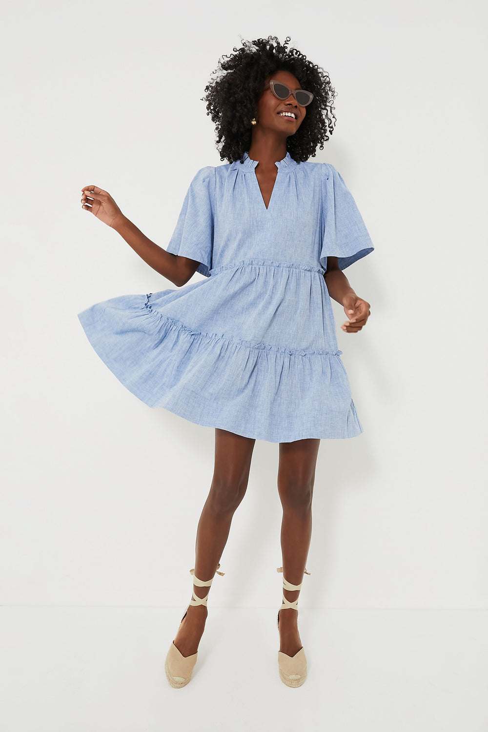 A woman in a Tuckernuck light blue Chambray Crawford Dress with flutter sleeves, ruffle detailing, and beige espadrille wedges smiles and strikes a playful pose.