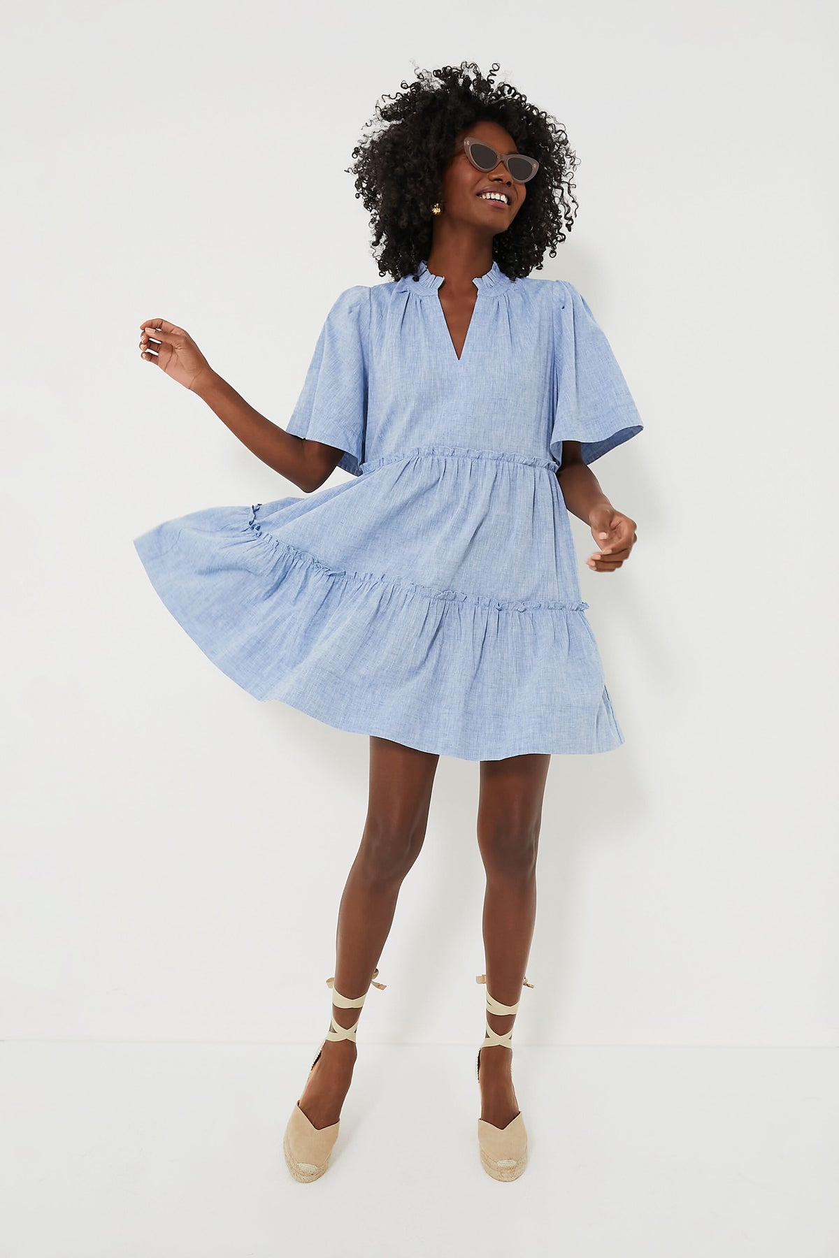 A woman in a Tuckernuck light blue Chambray Crawford Dress with flutter sleeves, ruffle detailing, and beige espadrille wedges smiles and strikes a playful pose.
