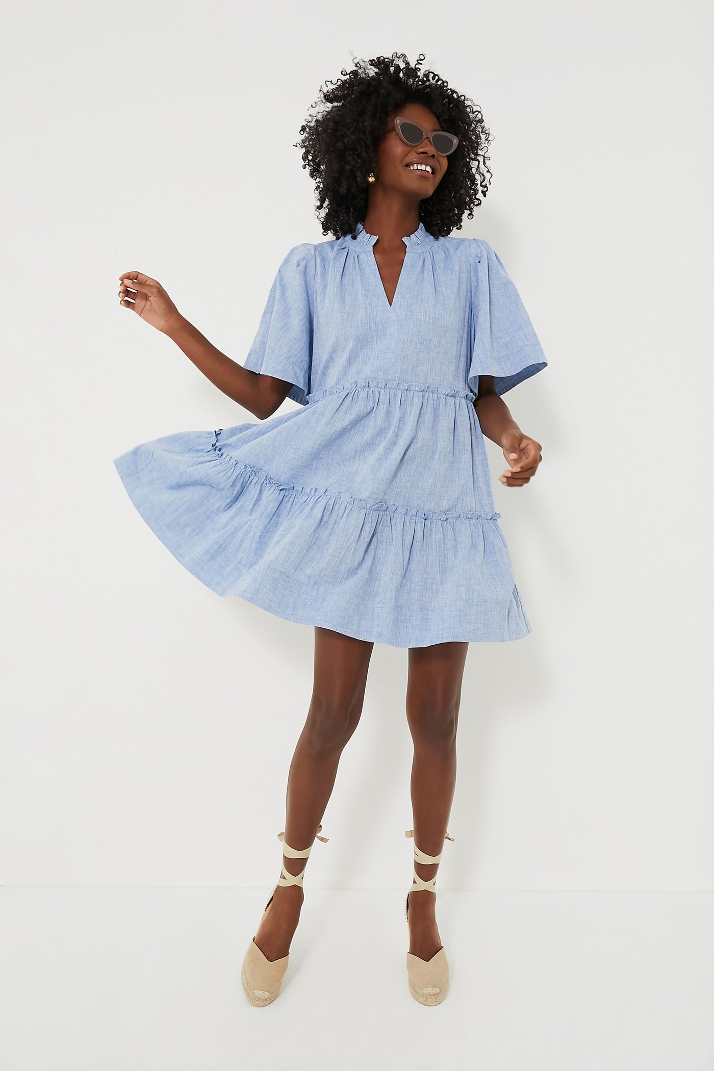 A woman in a Tuckernuck light blue Chambray Crawford Dress with flutter sleeves, ruffle detailing, and beige espadrille wedges smiles and strikes a playful pose.