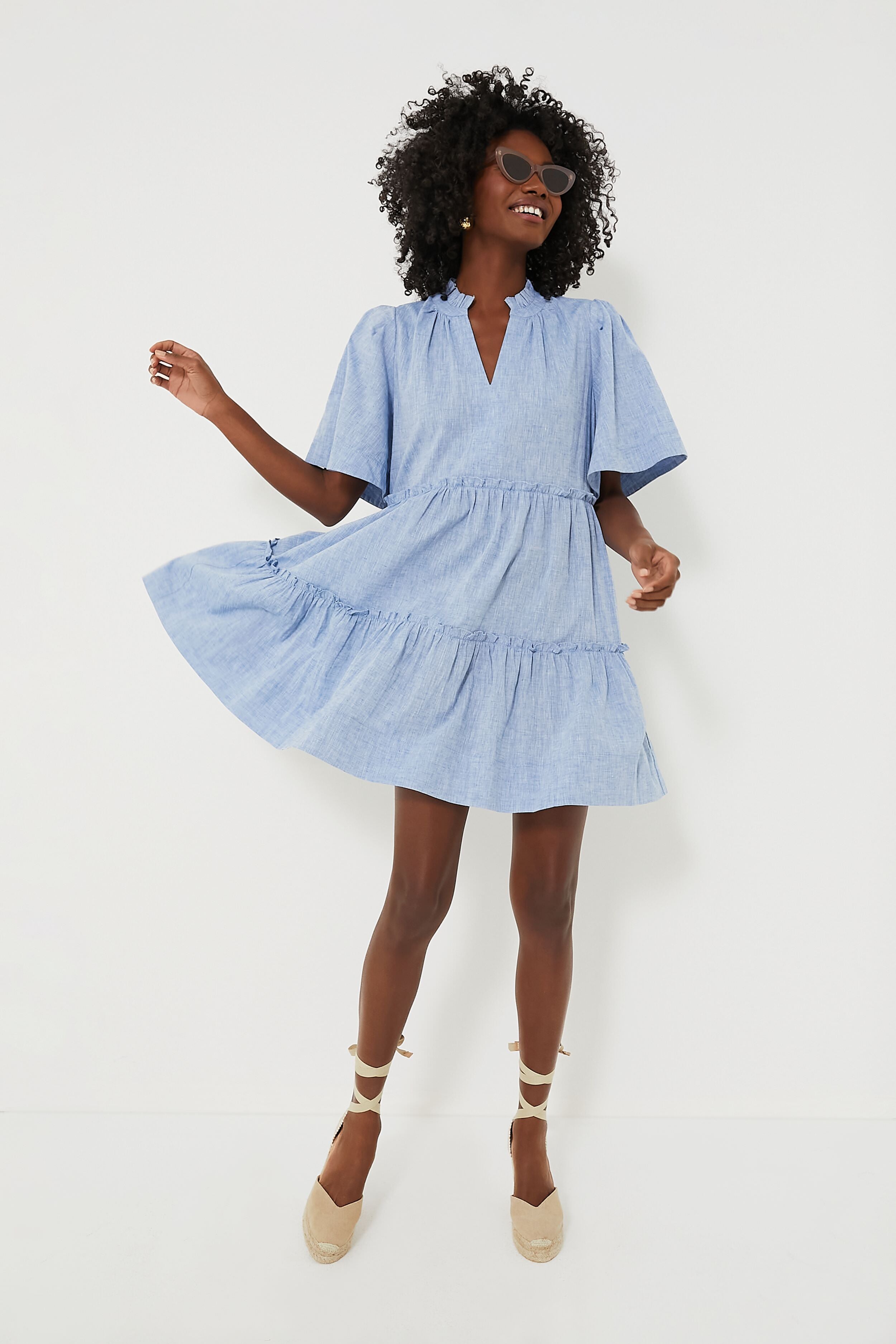 A woman in a Tuckernuck light blue Chambray Crawford Dress with flutter sleeves, ruffle detailing, and beige espadrille wedges smiles and strikes a playful pose.
