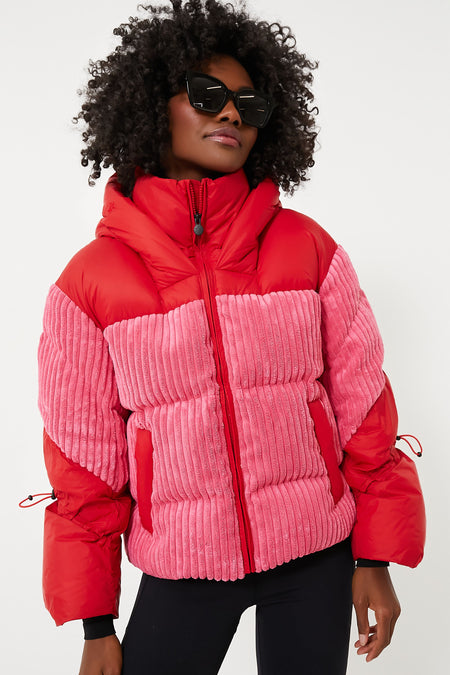 A person confidently stands wearing a Perfect Moment red and pink down jacket with corduroy panels, black leggings, and large black sunglasses, showcasing high-performance lifestyle clothing.