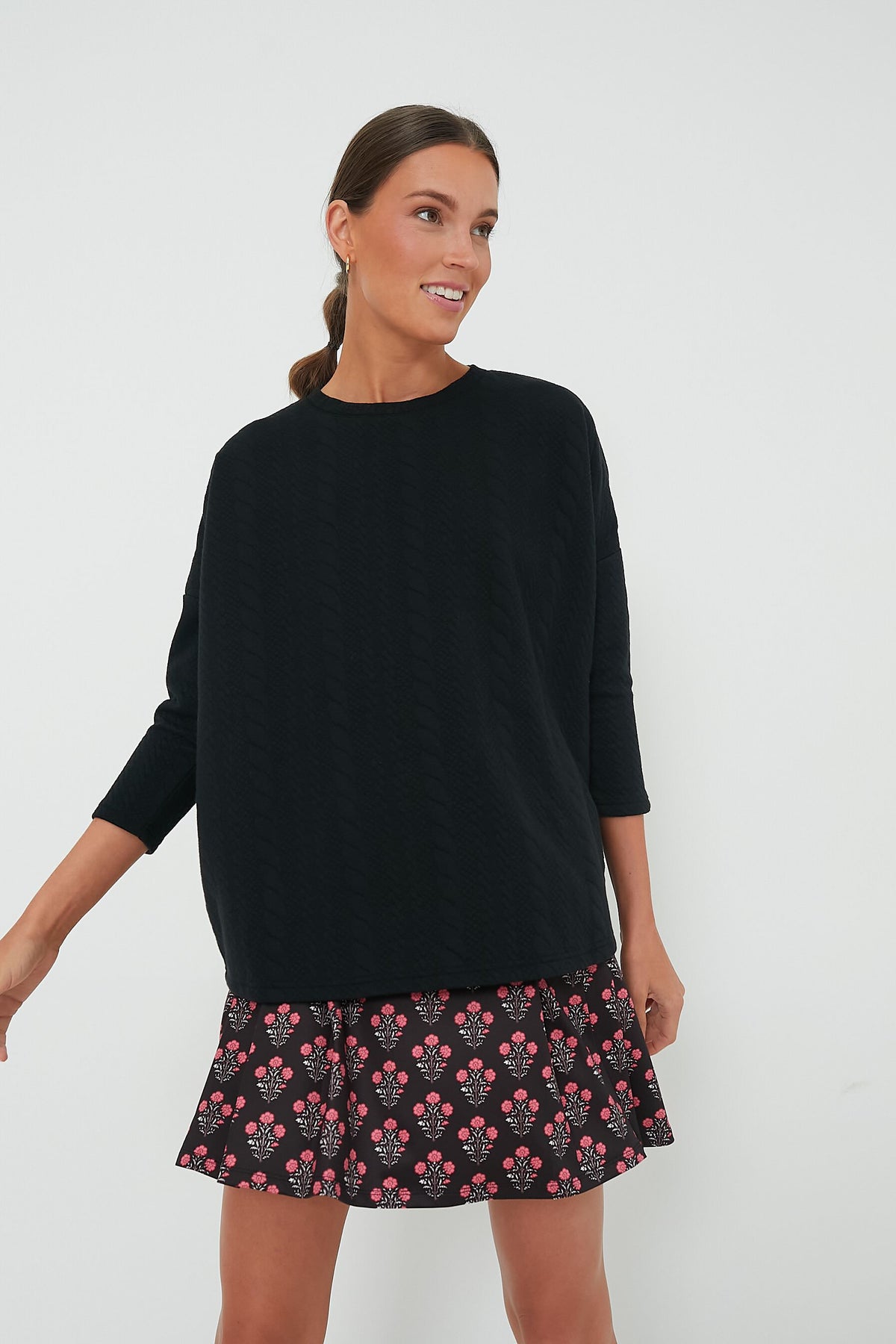 A woman wearing a relaxed fit Tnuck Sport sweatshirt and a short black skirt with a pink floral pattern, looking to the side and smiling.