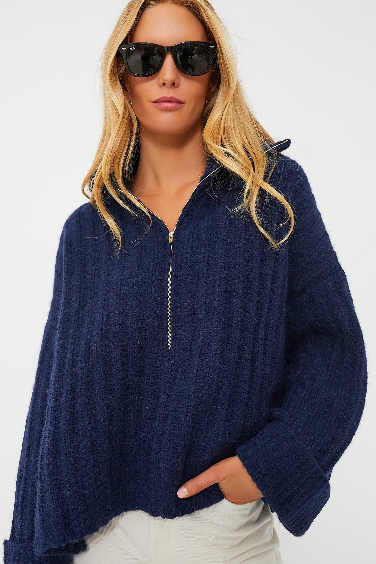 A woman models ELEVEN SIX's navy blue oversized sweater with Andean-inspired knitwork, paired with black sunglasses and light-colored pants, her hand in the pocket.
