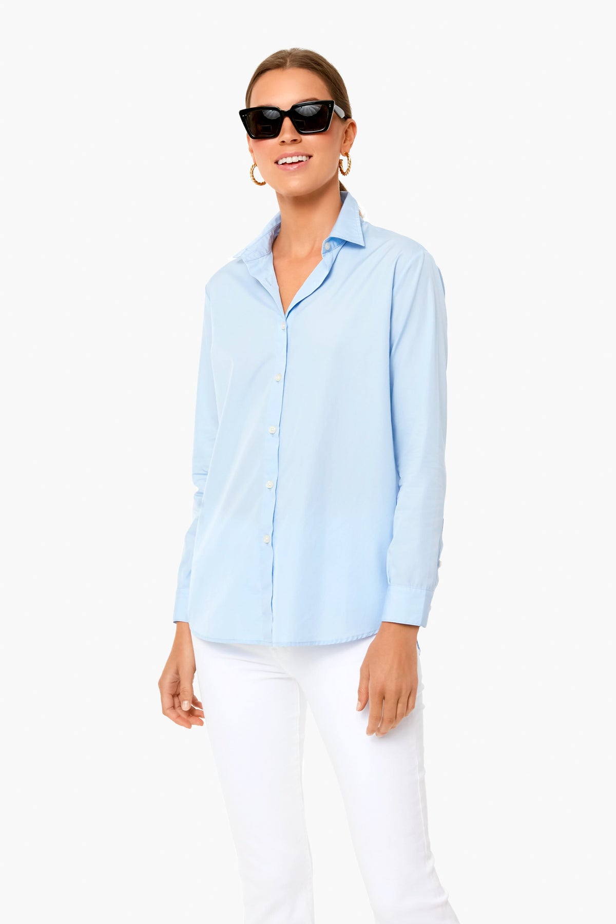 A woman in black sunglasses, gold hoop earrings, a KULE Italian cotton button-down shirt, and white pants stands confidently.