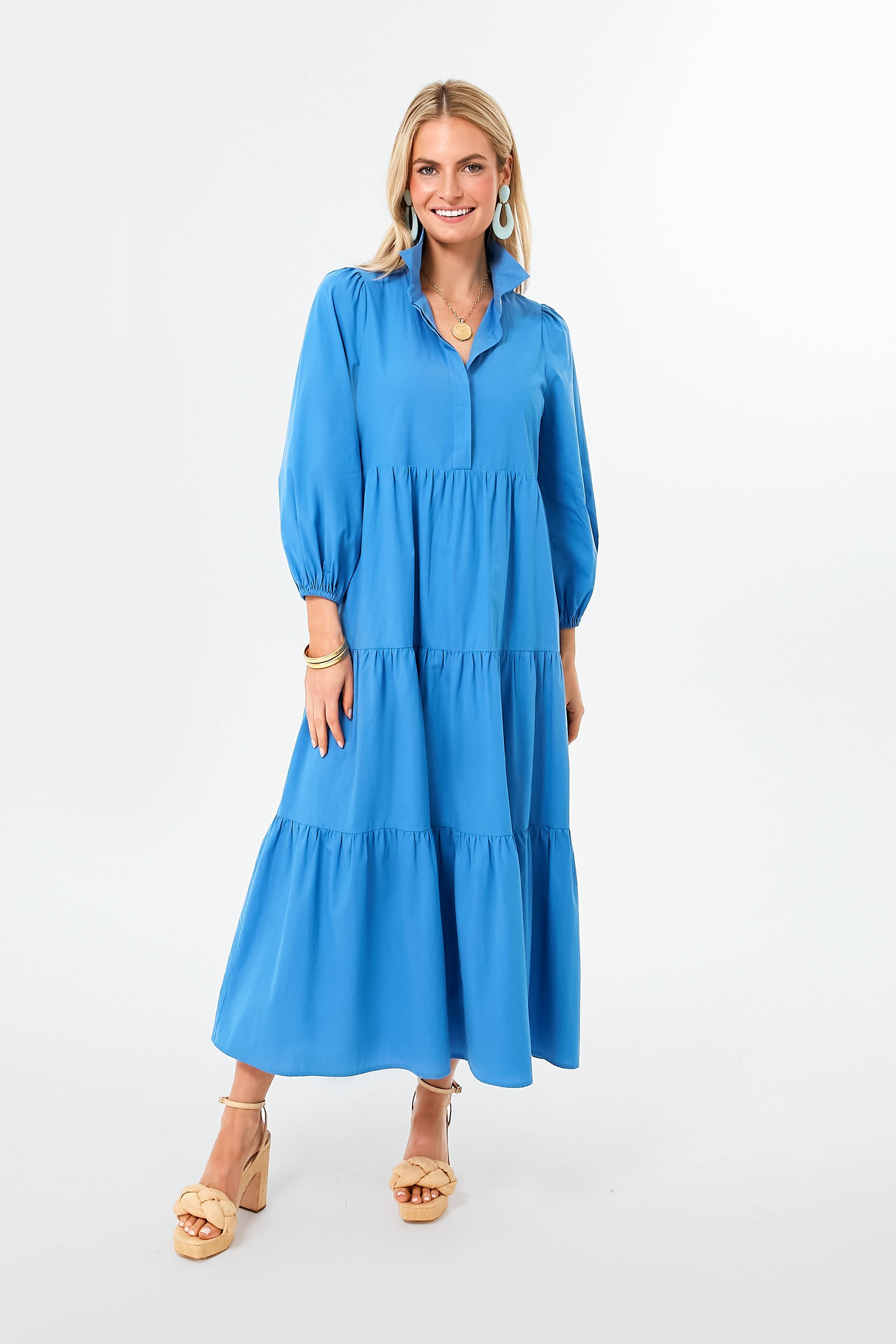 A woman stands wearing a Pomander Place long-sleeved blue tiered maxi dress with puff sleeves, tan platform sandals, and blue earrings.